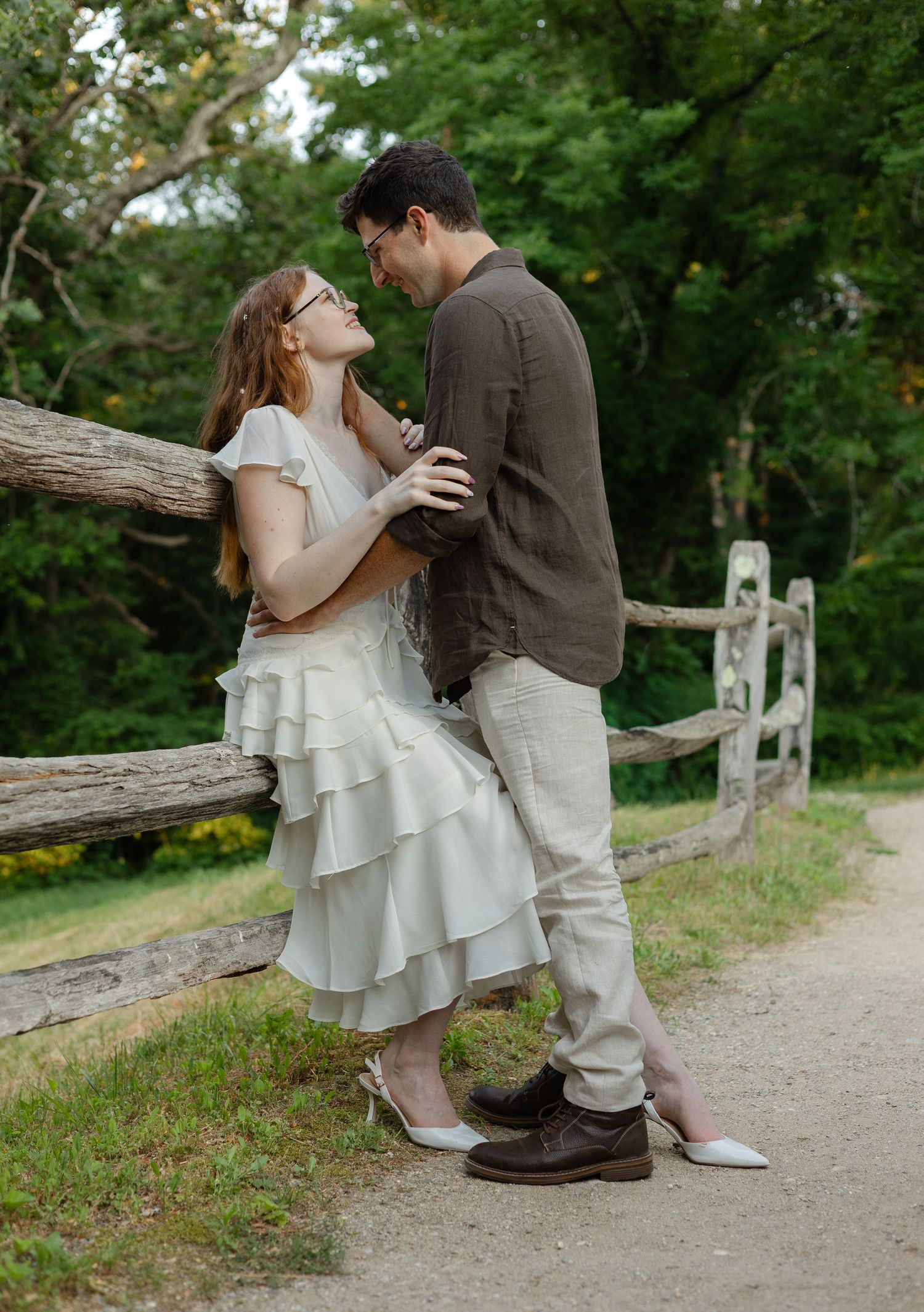 A couple shares a romantic moment by a rustic wooden fence, woman in white ruffled dress leaning toward man in brown shirt.