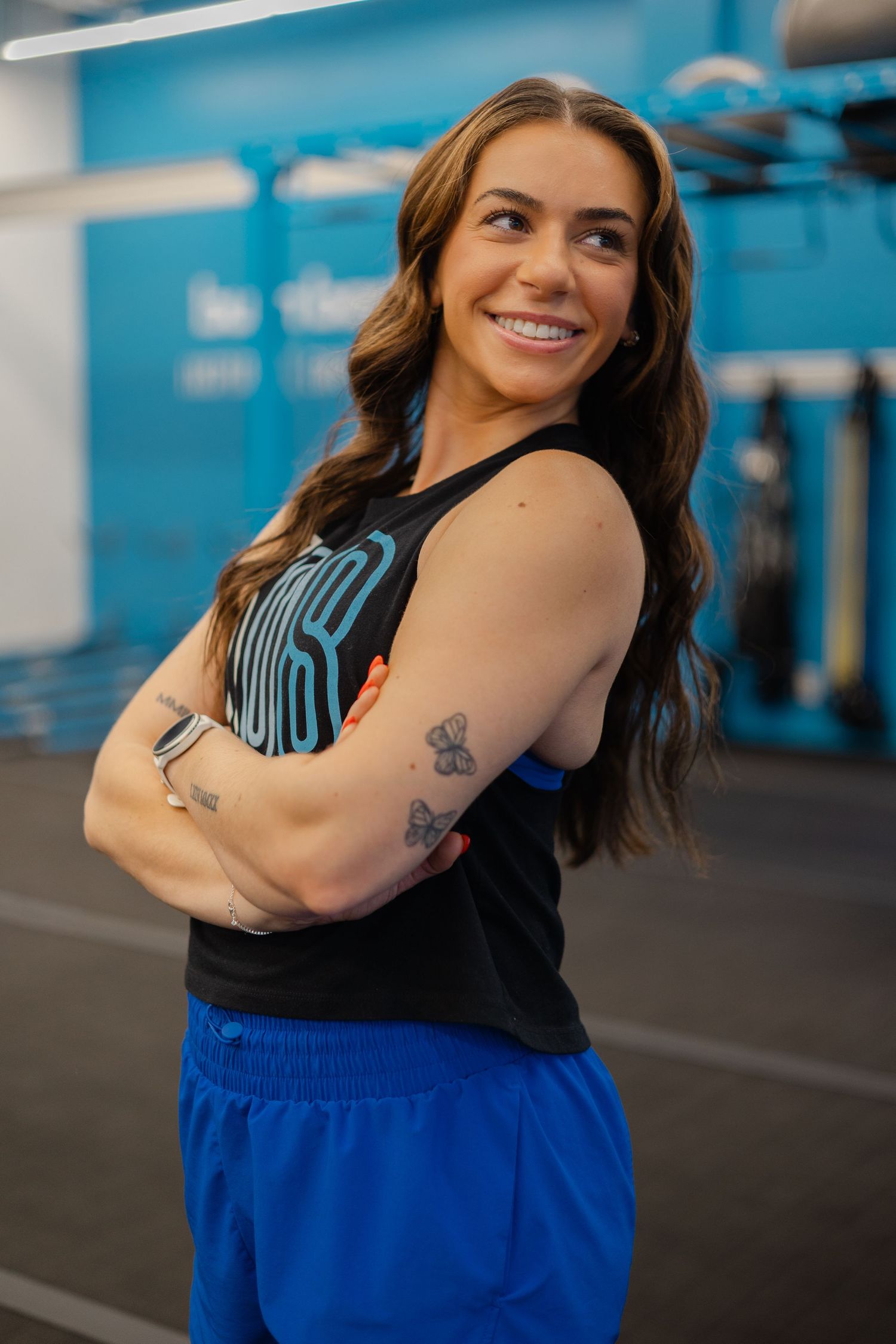 Smiling woman in black tank top and blue shorts stands with arms crossed in a gym setting.