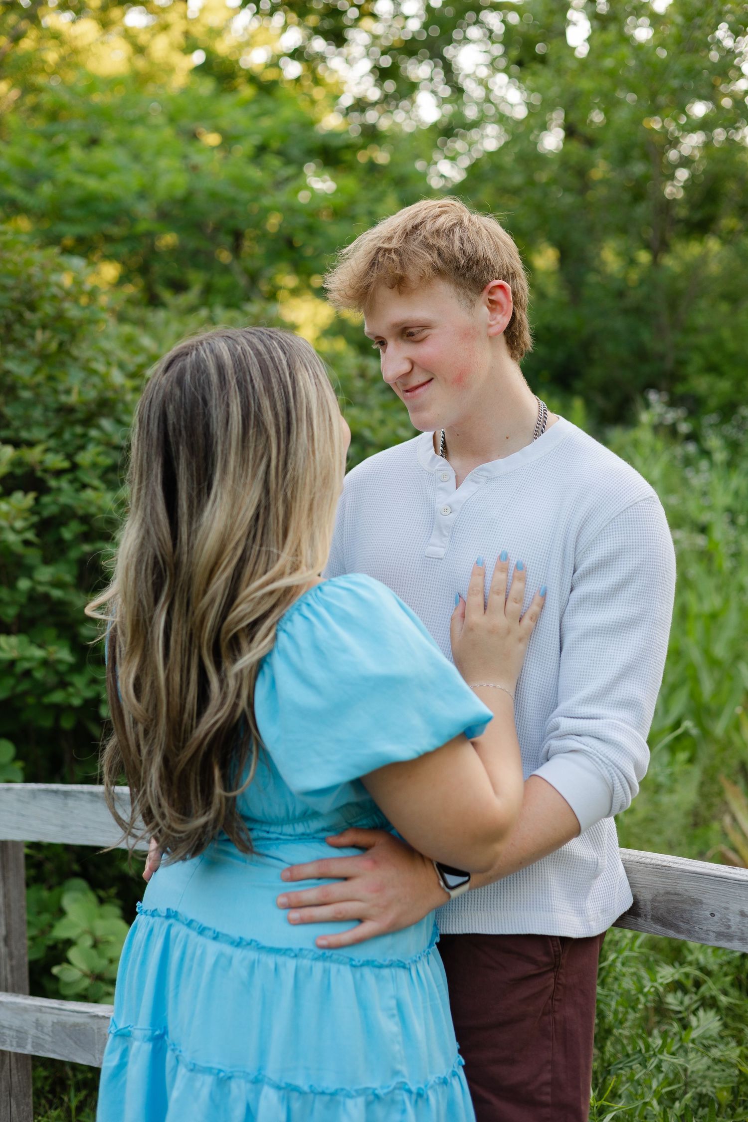 Young couple embracing outdoors near wooden fence, woman in blue dress facing man in white shirt surrounded by greenery.