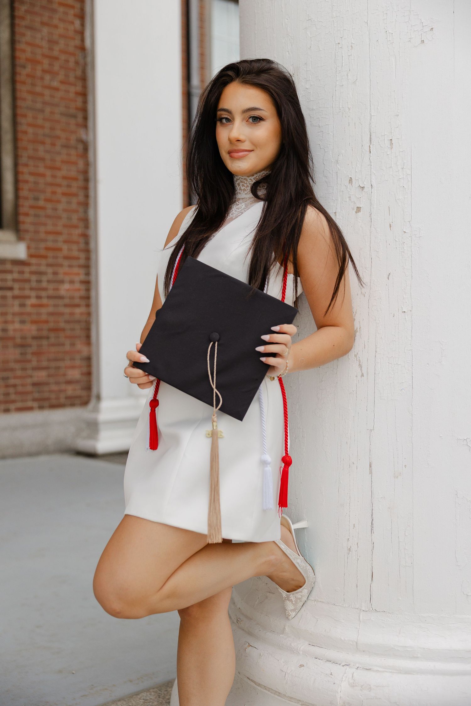 Brunette graduate in white mini dress holding black cap with red tassel, leaning against white column.