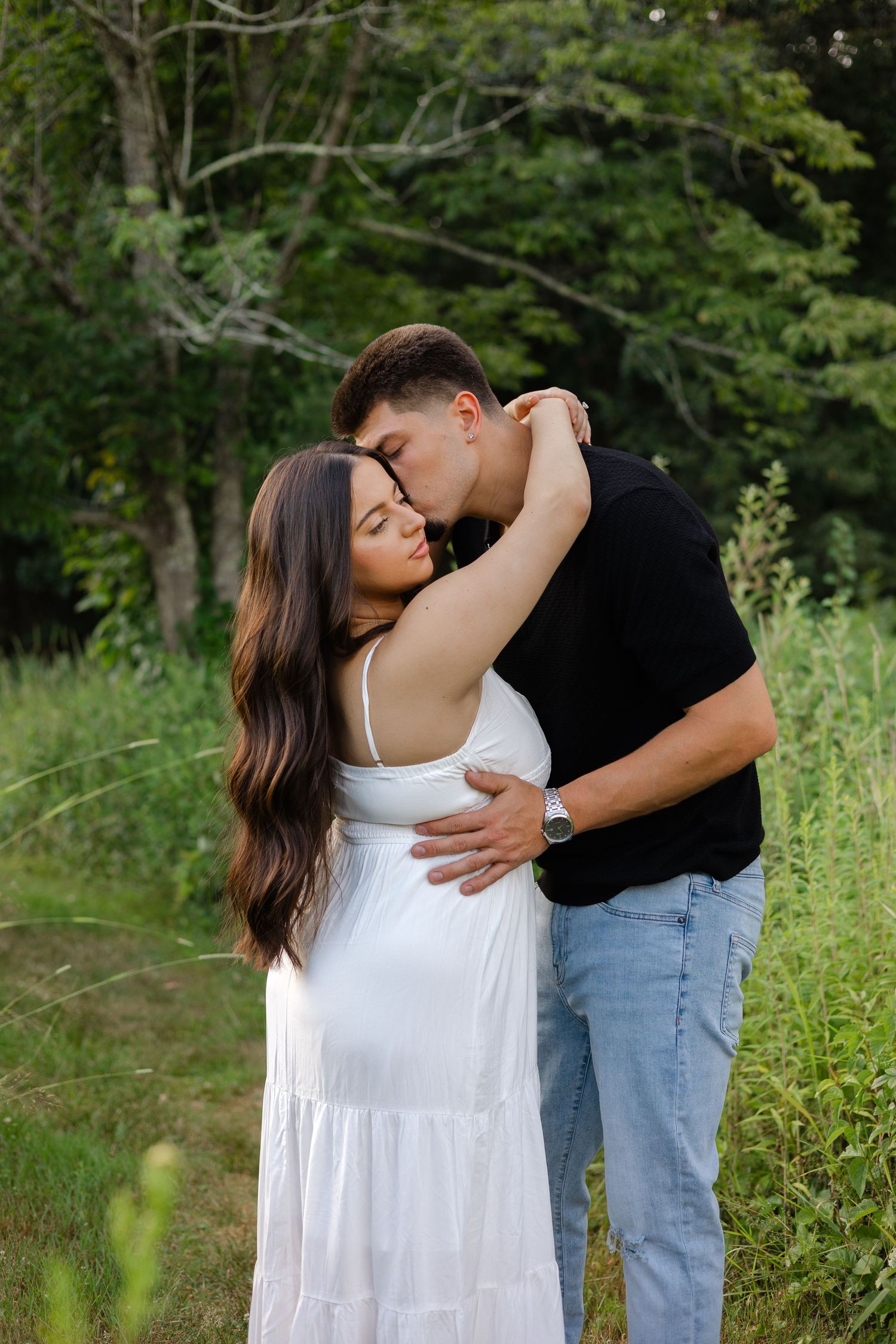 A couple shares a tender embrace outdoors, man in black shirt kissing woman in white dress amid lush green trees.