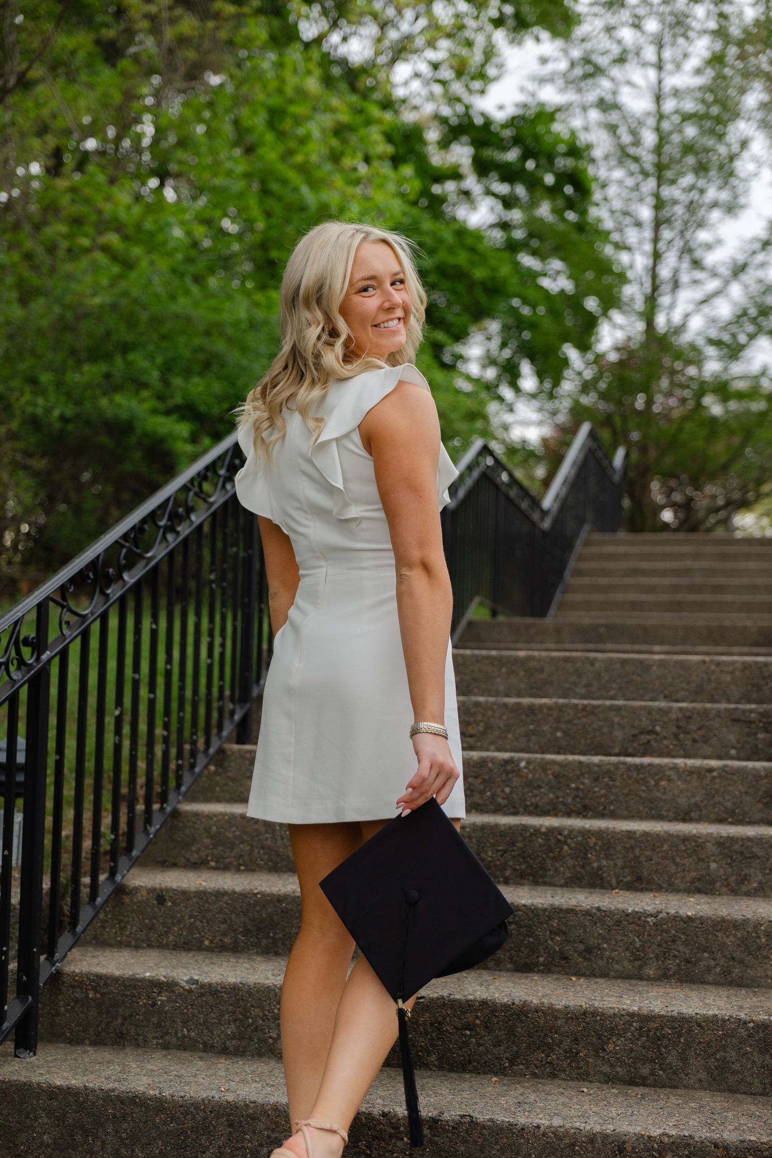 Blonde graduate in white dress carrying black cap while walking up stone staircase outdoors.