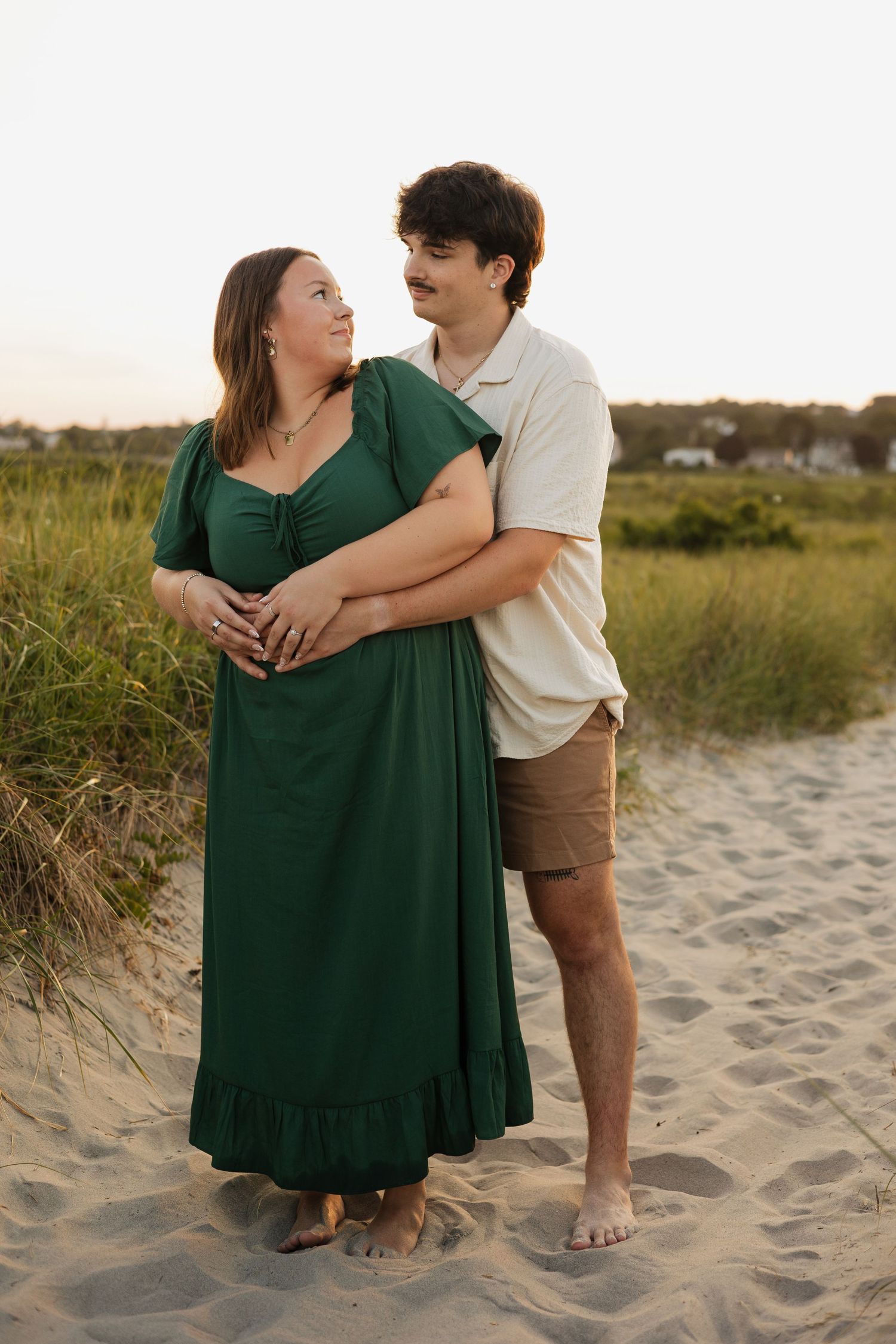 Couple embracing on sandy beach, woman in green dress, man in cream shirt and khaki shorts, at sunset.