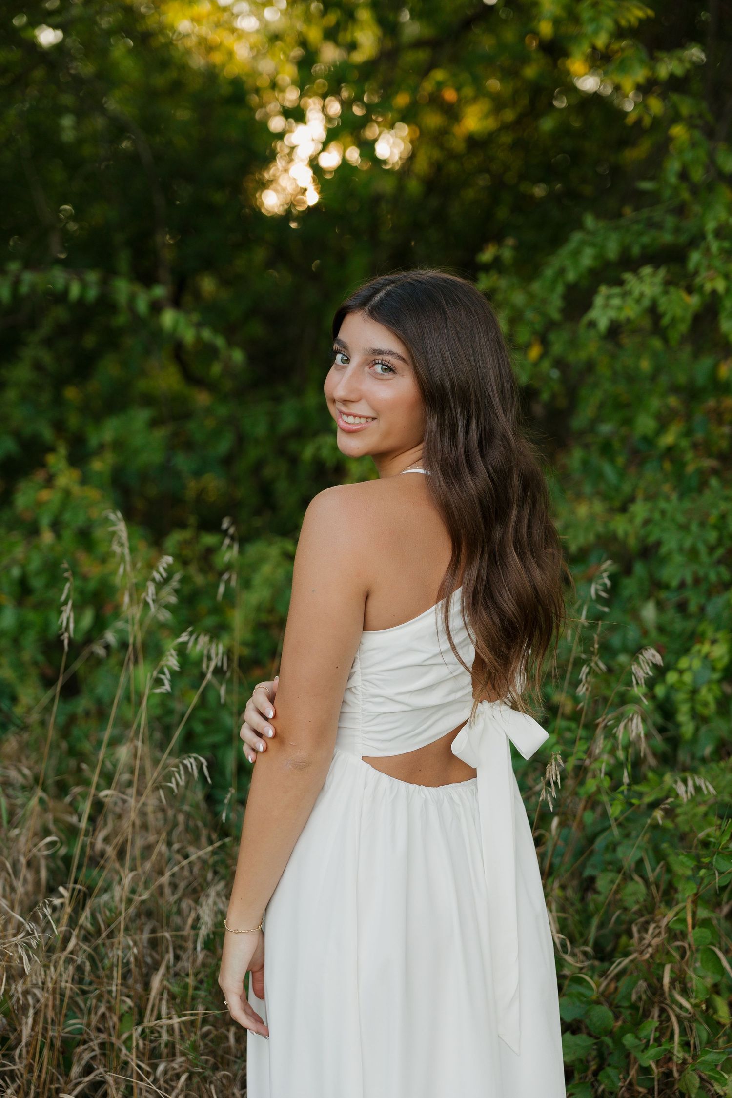 Young woman in white strapless dress smiling over shoulder in golden hour outdoor setting.