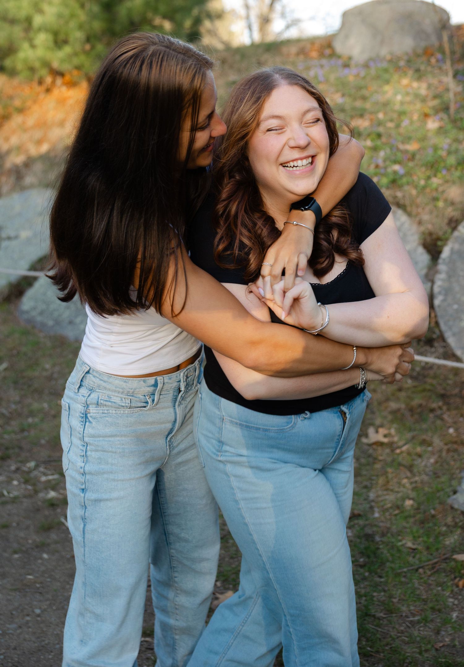 Two young women hugging and laughing together outdoors near large rocks in warm sunlight.