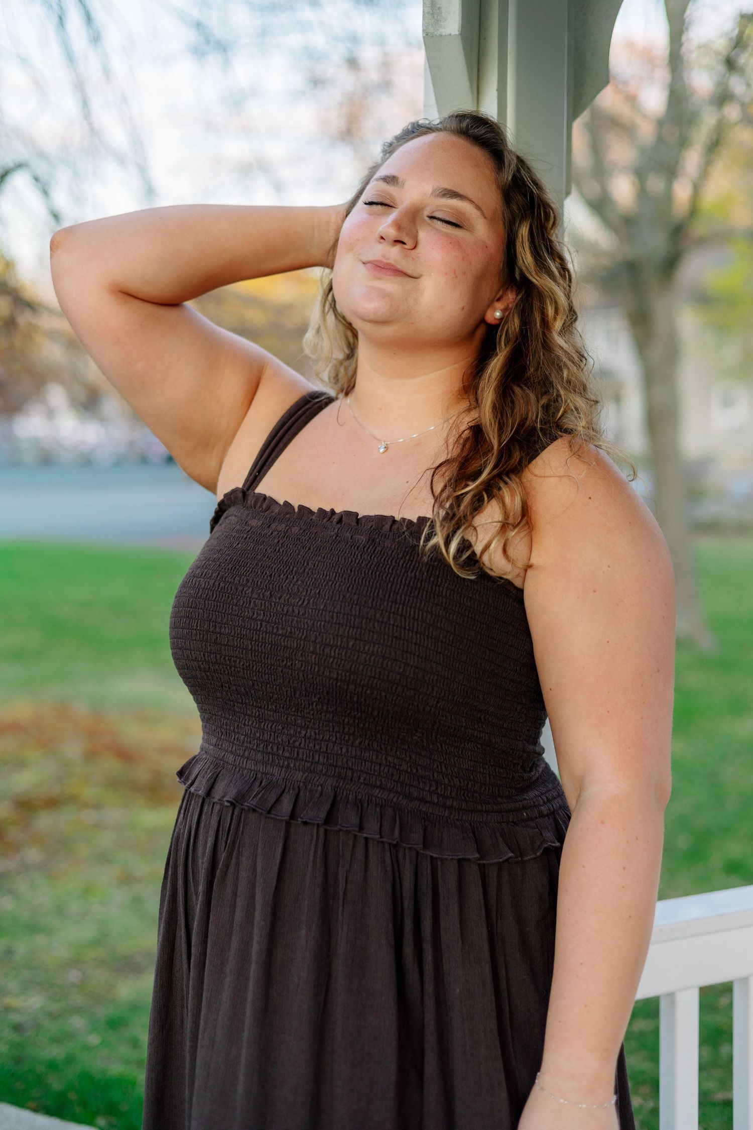 Young woman in black smocked sundress posing on porch with hand on neck, eyes closed, in soft natural light.