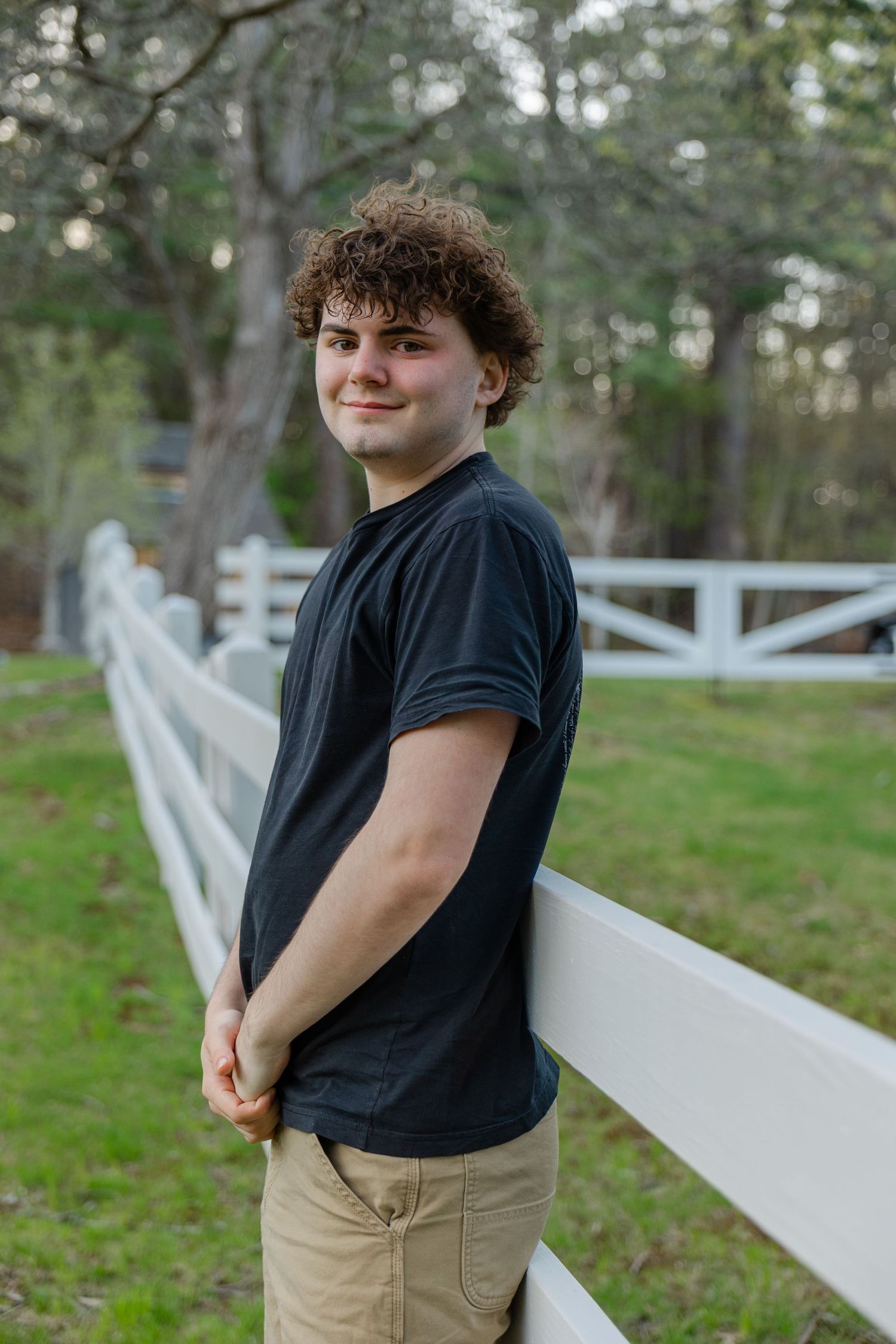 Smiling curly-haired teen boy leaning against white fence in park, wearing black tee and khaki pants.
