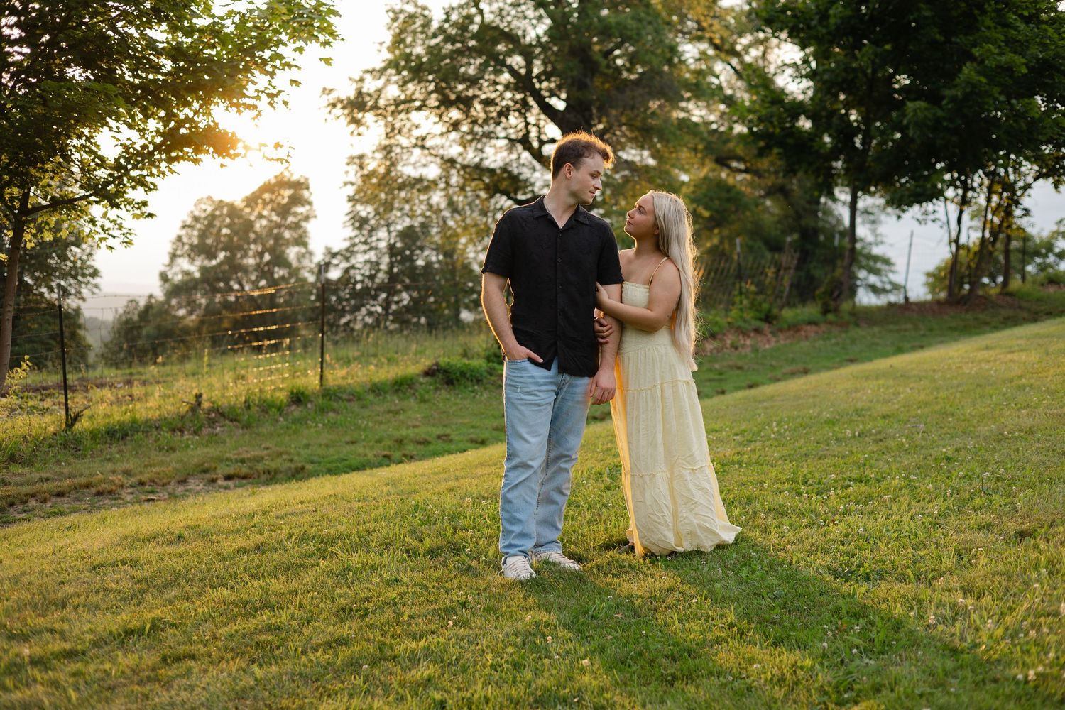 Couple holding hands in a sunlit green field at golden hour, woman in cream dress, man in black shirt and jeans.