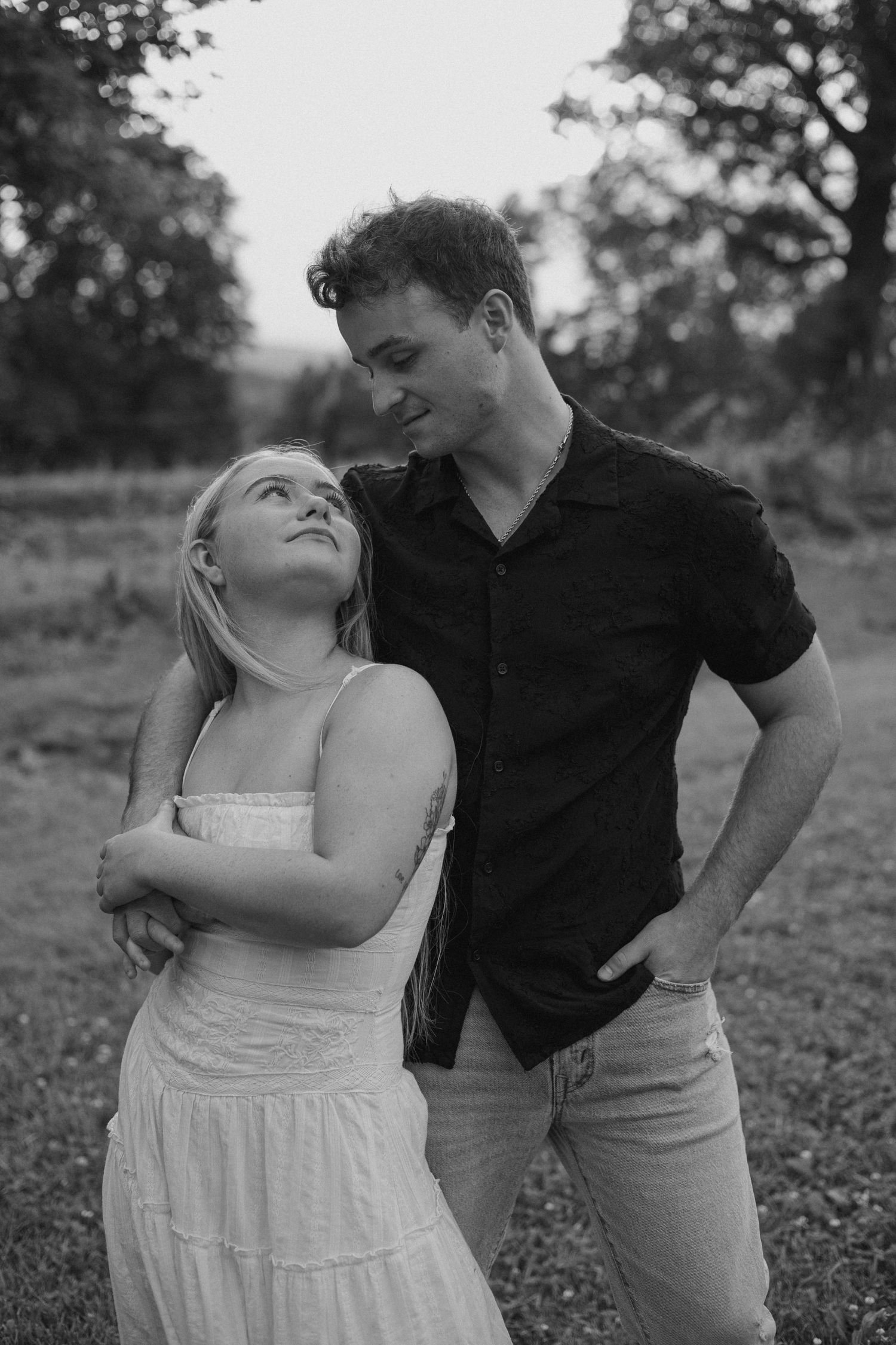 Black and white photo of a young couple sharing a tender moment outdoors, woman in white dress looking up at man in black shirt.