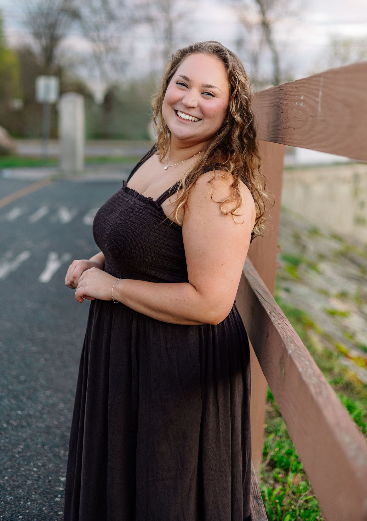 Young woman in black maxi sundress smiling while leaning against wooden fence on a paved path at dusk.