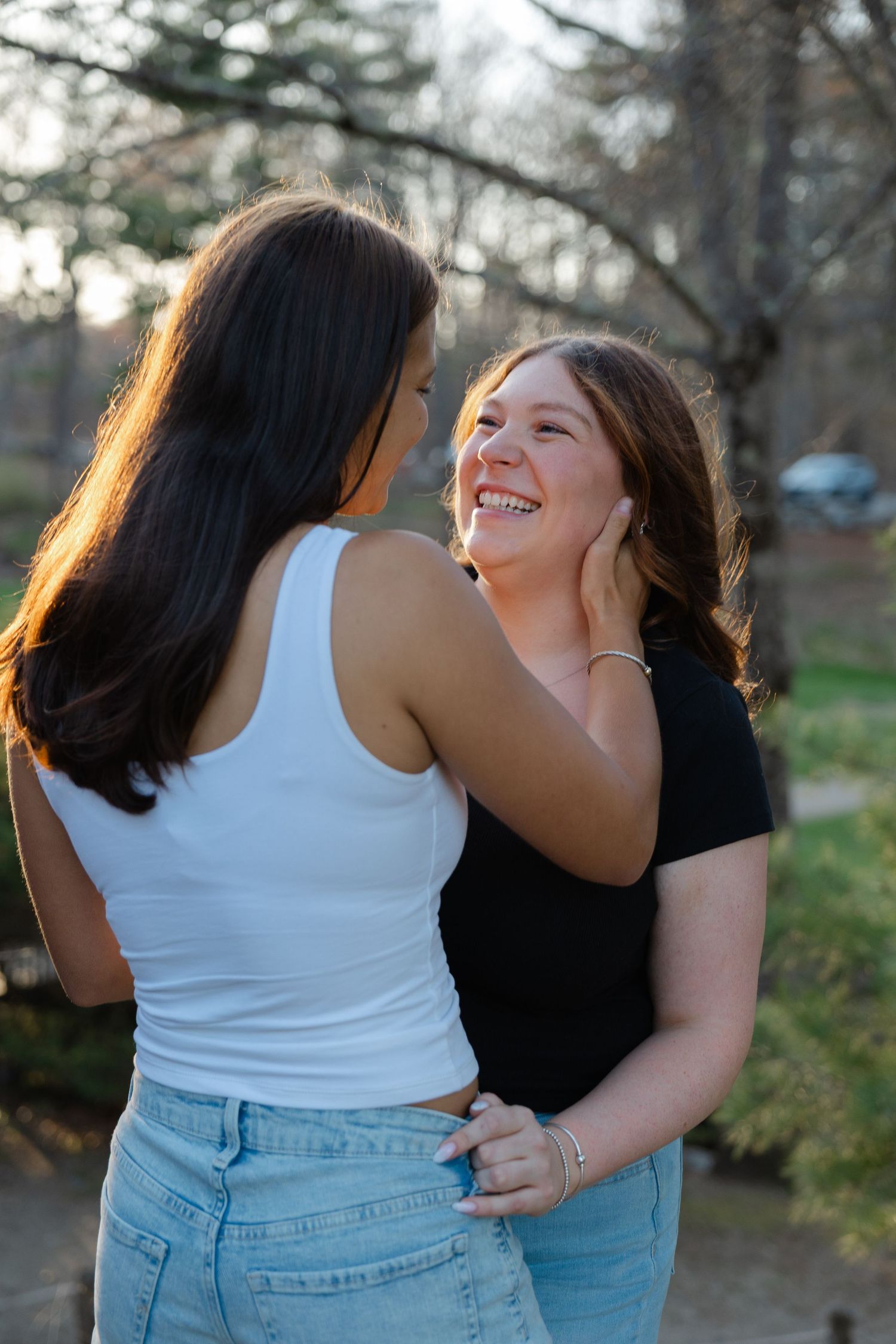 Two young women smiling and holding each other's faces tenderly outdoors.