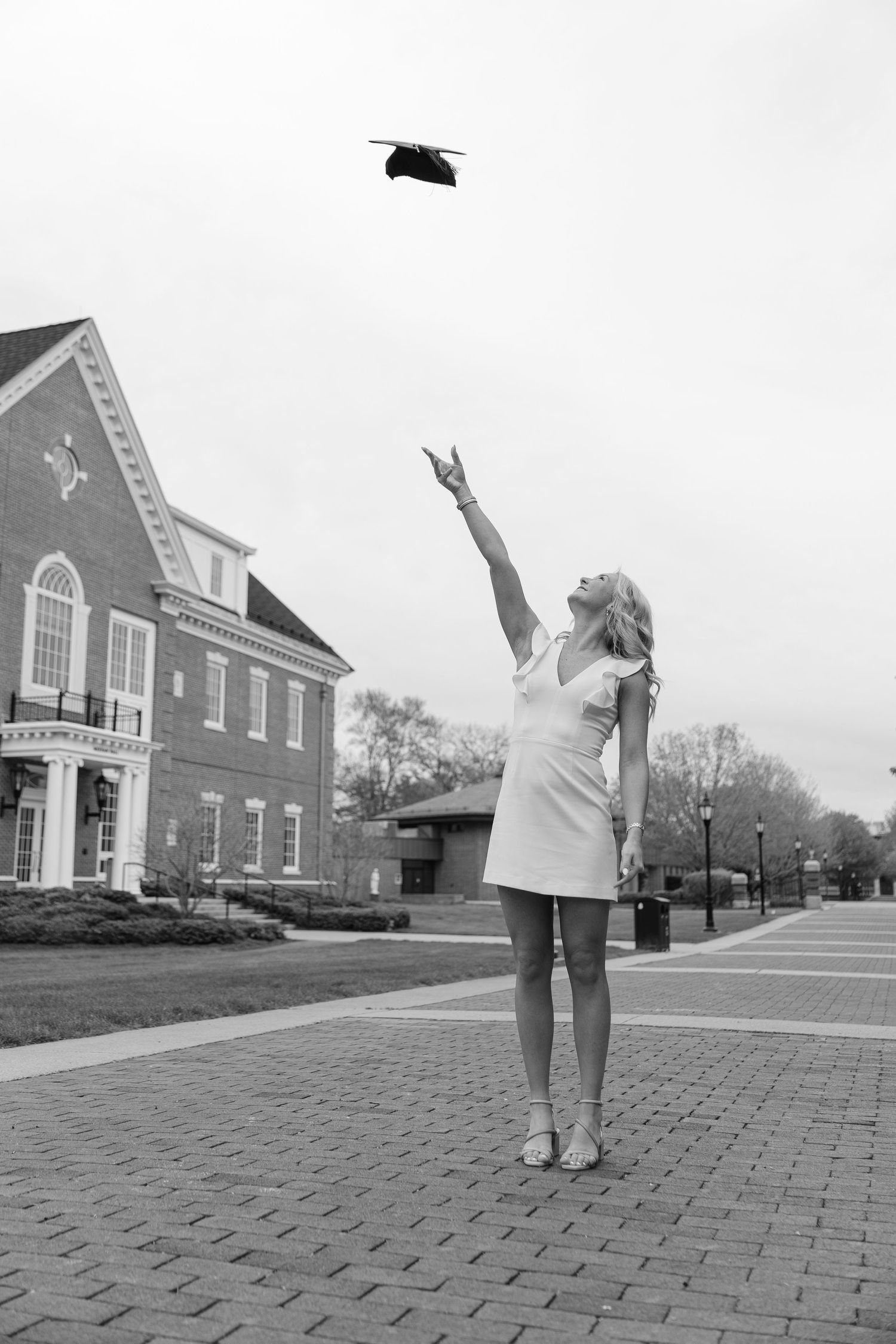 Black and white photo of a young woman in a dress tossing her graduation cap in the air on a brick campus pathway.