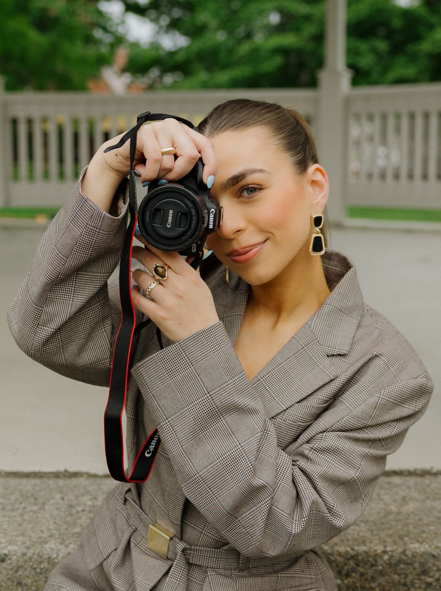 Female photographer in plaid belted blazer posing with Canon camera outdoors.
