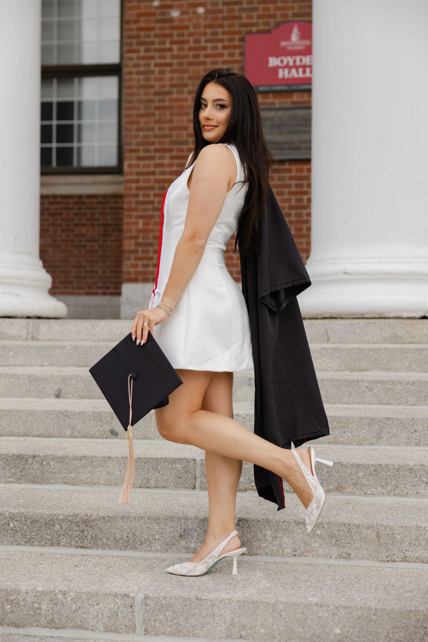 Elegant graduate in white dress and black gown holding cap on brick university building steps.