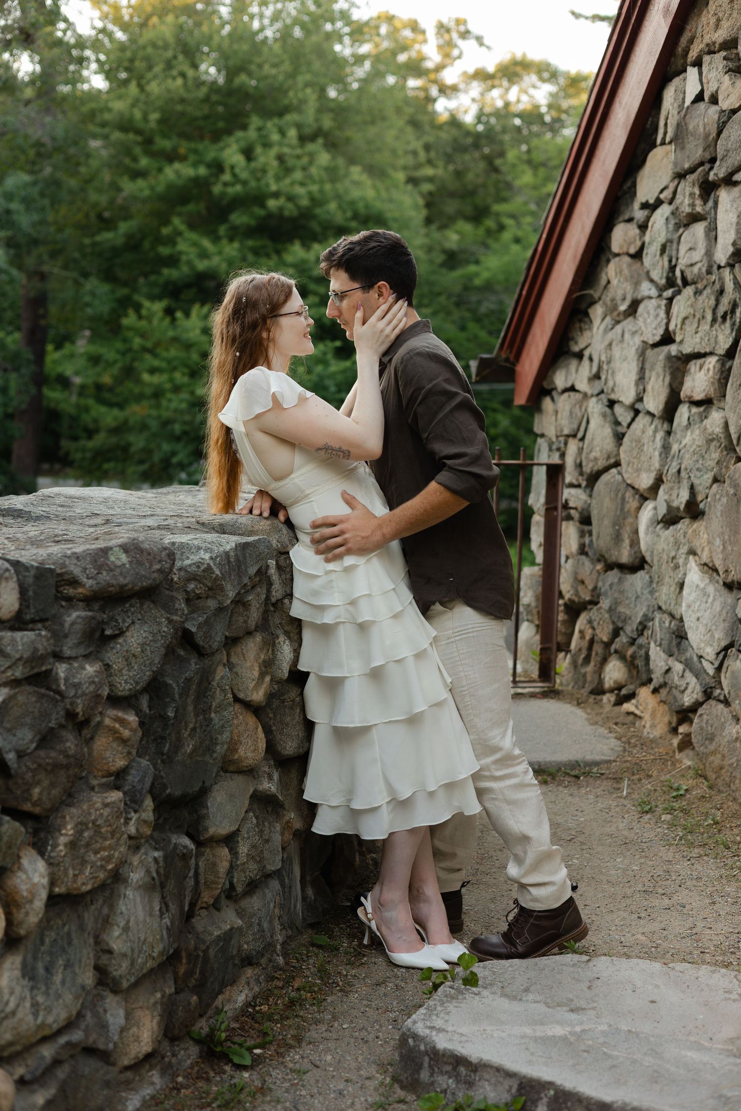 A couple shares a romantic moment near a stone wall, the woman in a white ruffled dress and the man in a brown jacket.