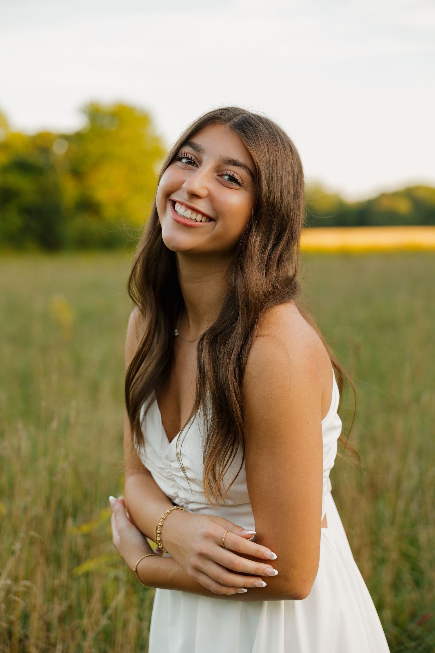 Young woman in white dress smiling in a green field at sunset, wearing gold bracelet.