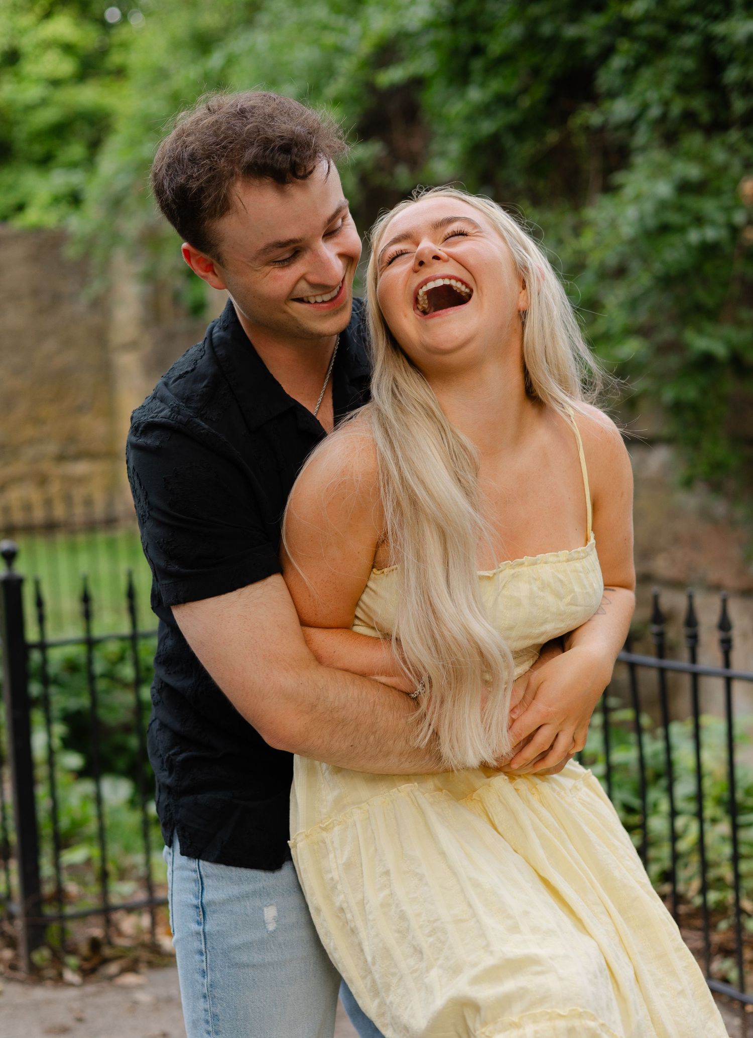 A joyful couple laughing and embracing outdoors, woman in yellow dress with long blonde hair, man in black shirt.
