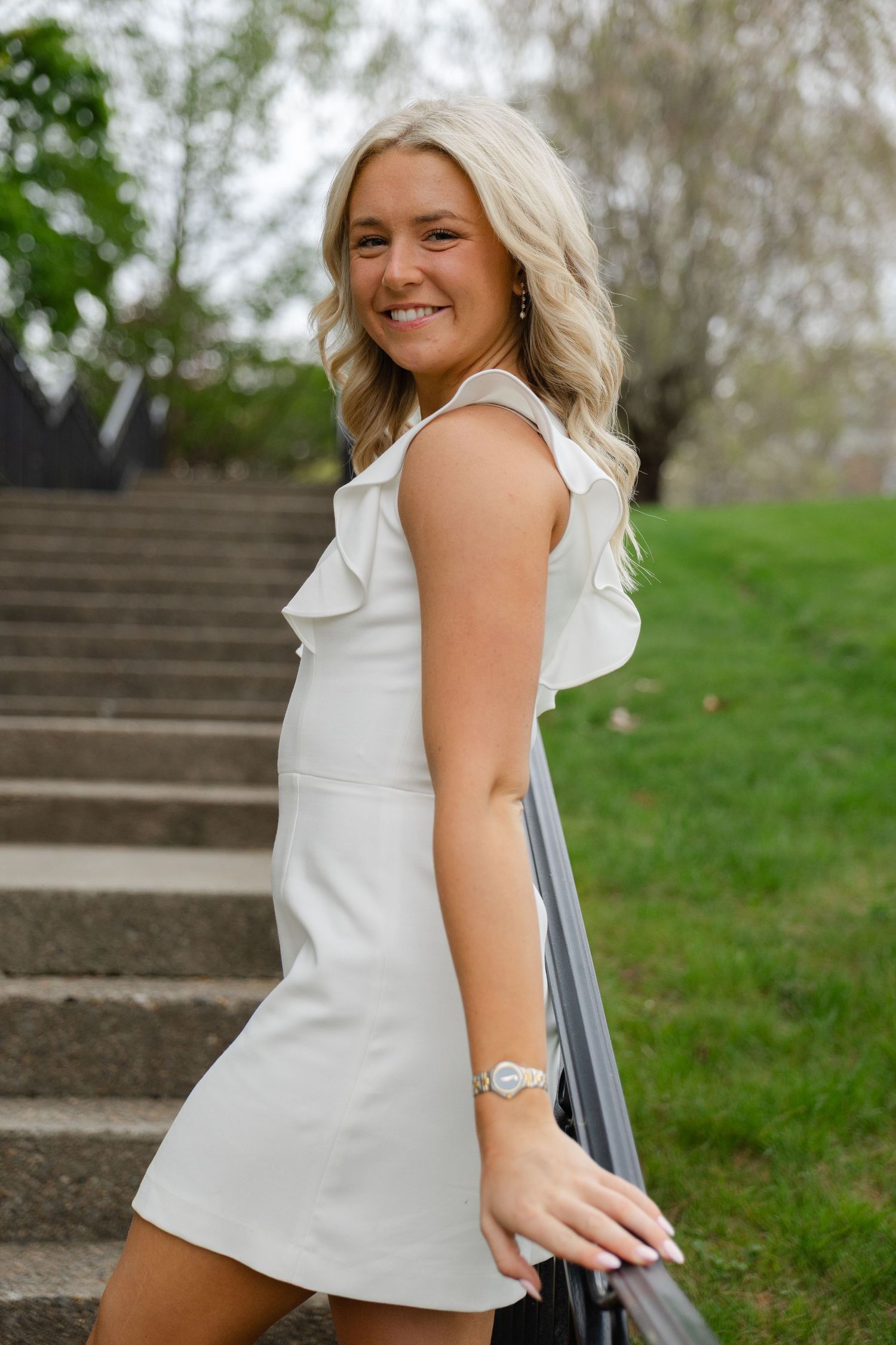 Smiling blonde graduate in white ruffled dress posing on outdoor campus staircase.