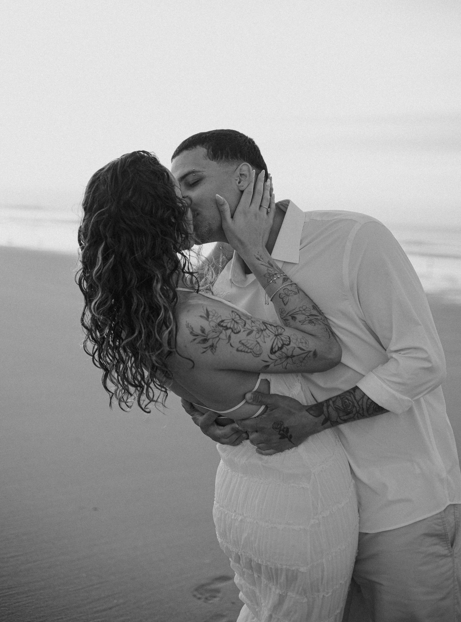 Black and white photo of tattooed couple sharing romantic kiss on beach at sunset.