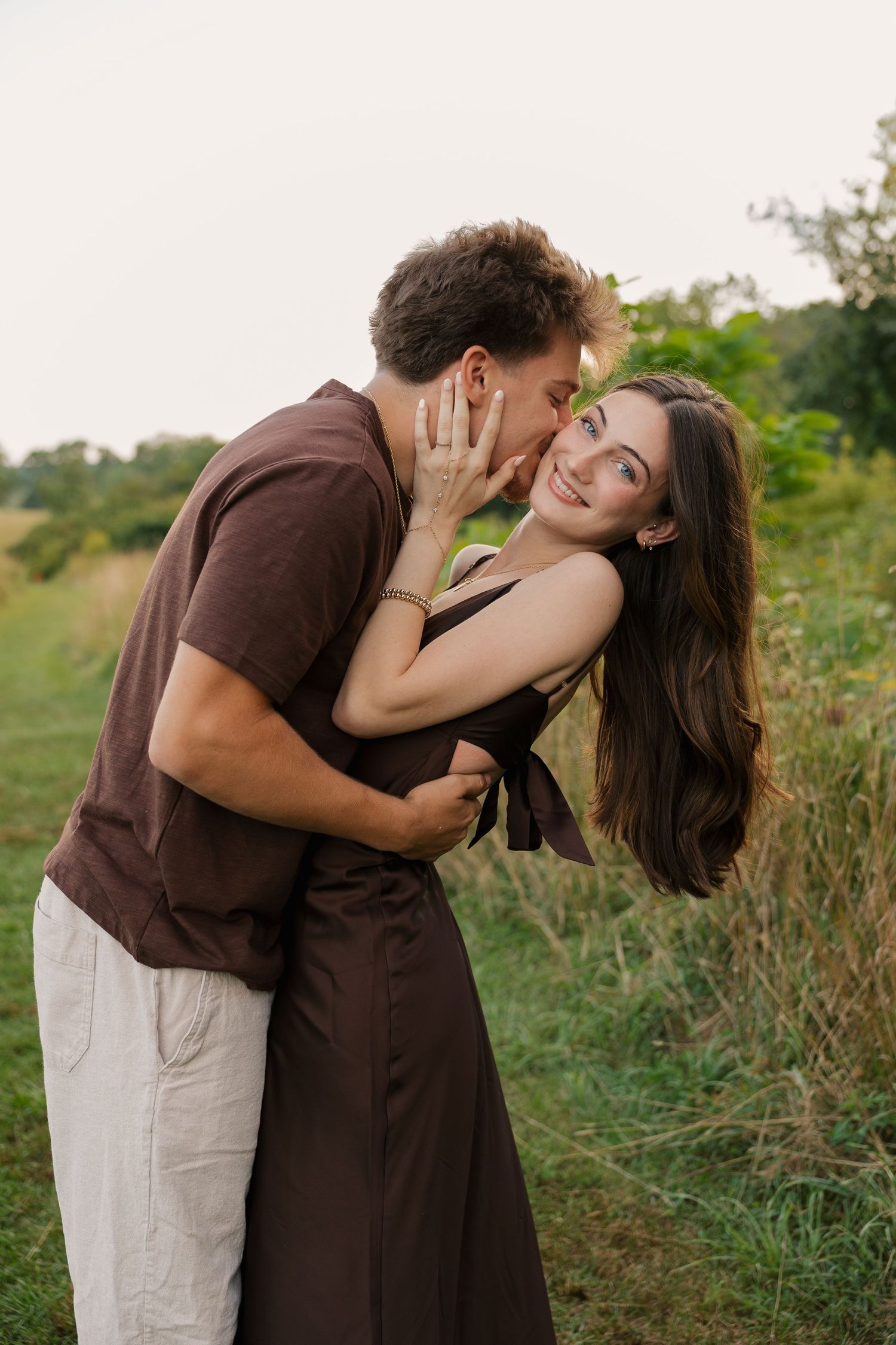 Romantic couple embracing outdoors, woman in brown dress smiling as man kisses her cheek in a green field.