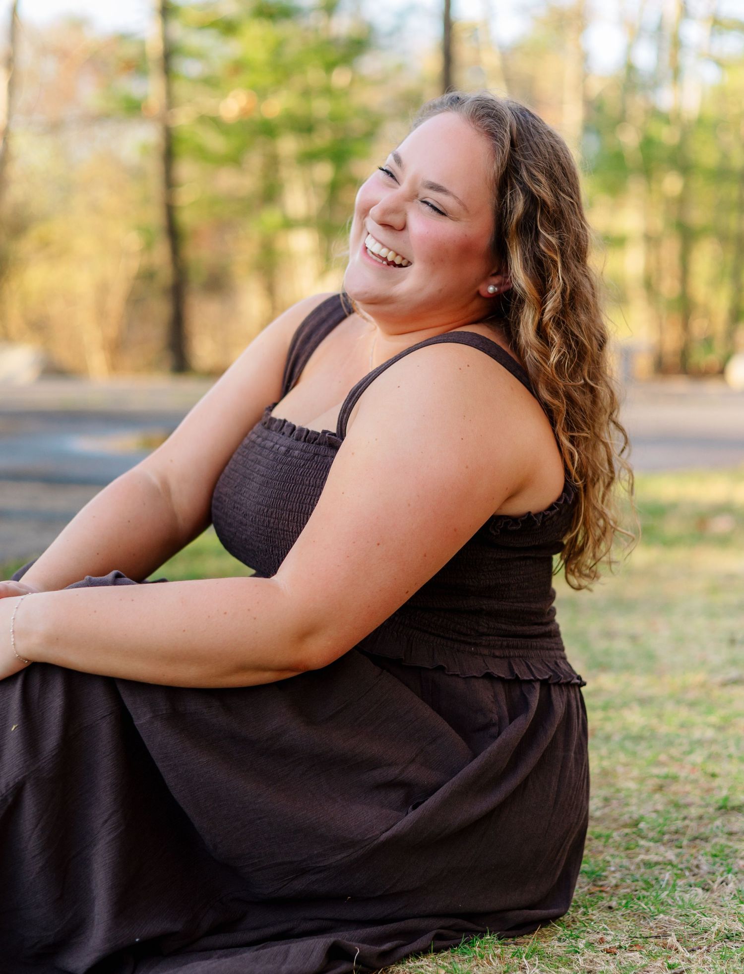 Young woman in black sundress sitting on grass laughing joyfully during golden hour outdoor photo session.
