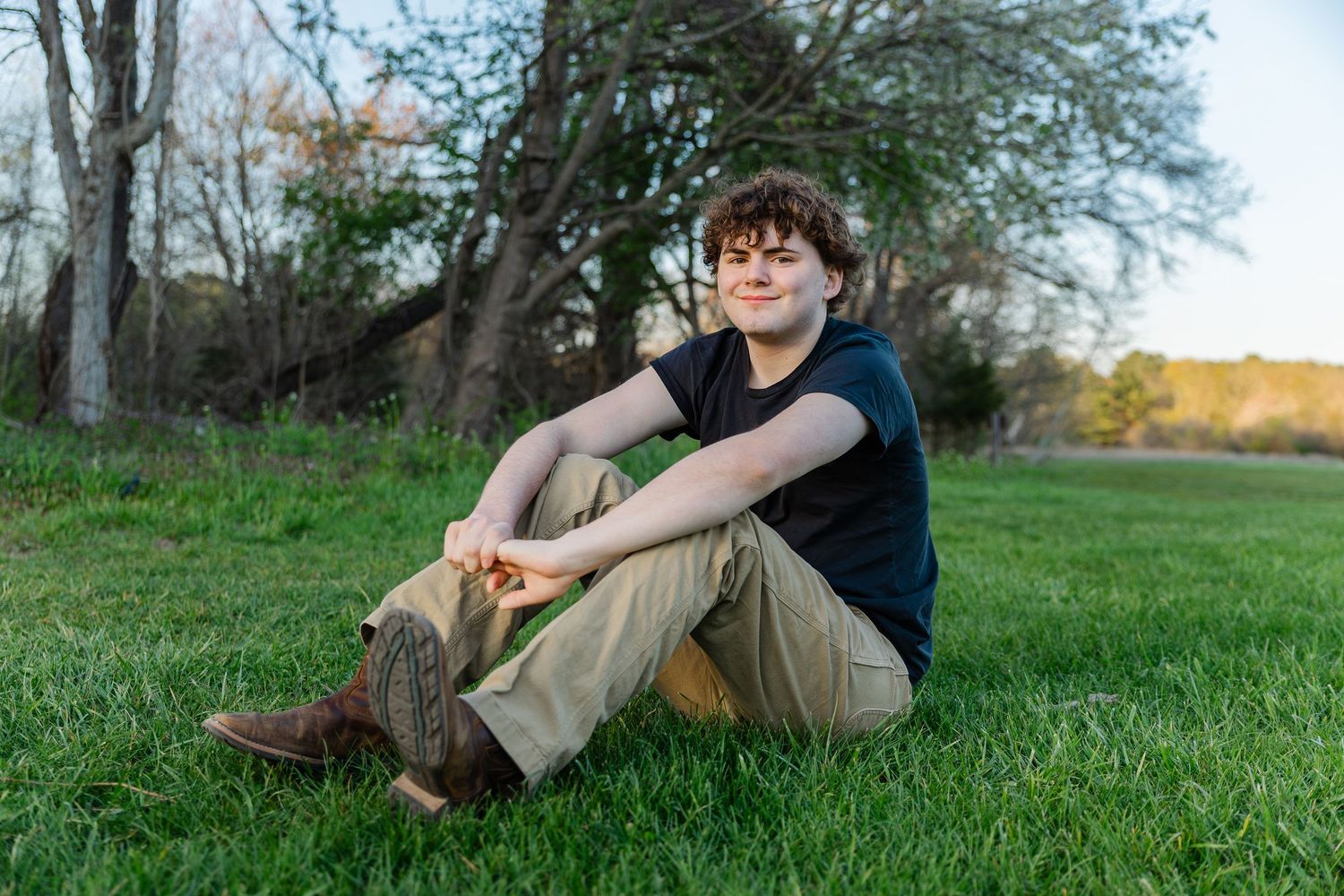 Teenage boy with curly hair sitting relaxed on green grass by tree, wearing black tee and khaki pants outdoors.