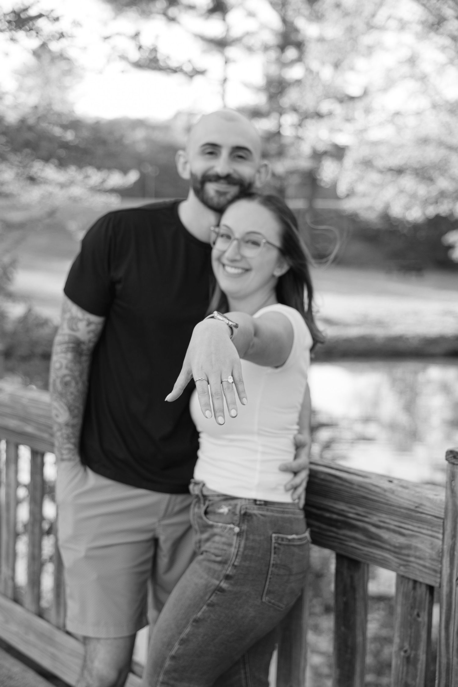 Black and white photo of engaged couple on wooden bridge, woman showing off engagement ring, smiling joyfully.