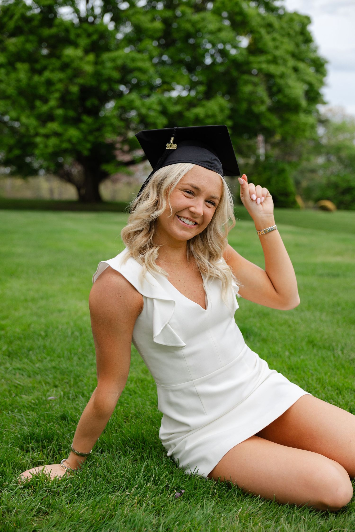 Smiling blonde graduate in white dress sitting on green grass, holding black graduation cap outdoors.