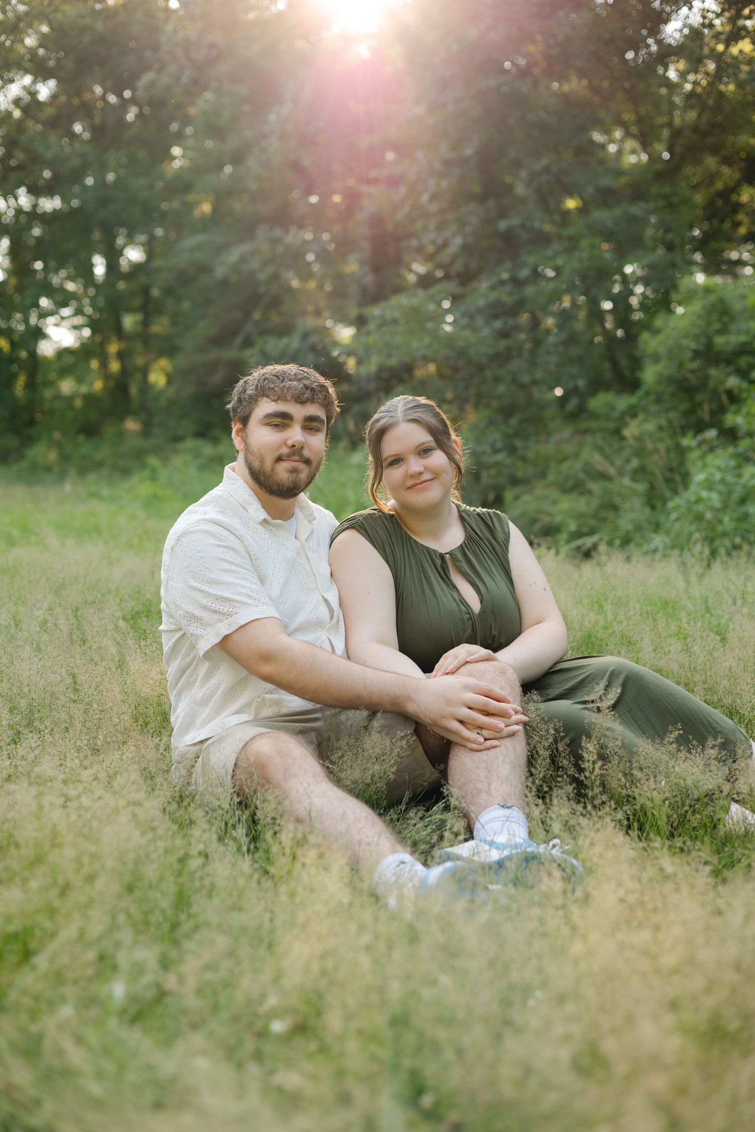 Couple sitting together in a sunlit grassy field, smiling, with lush green trees in the background during golden hour.