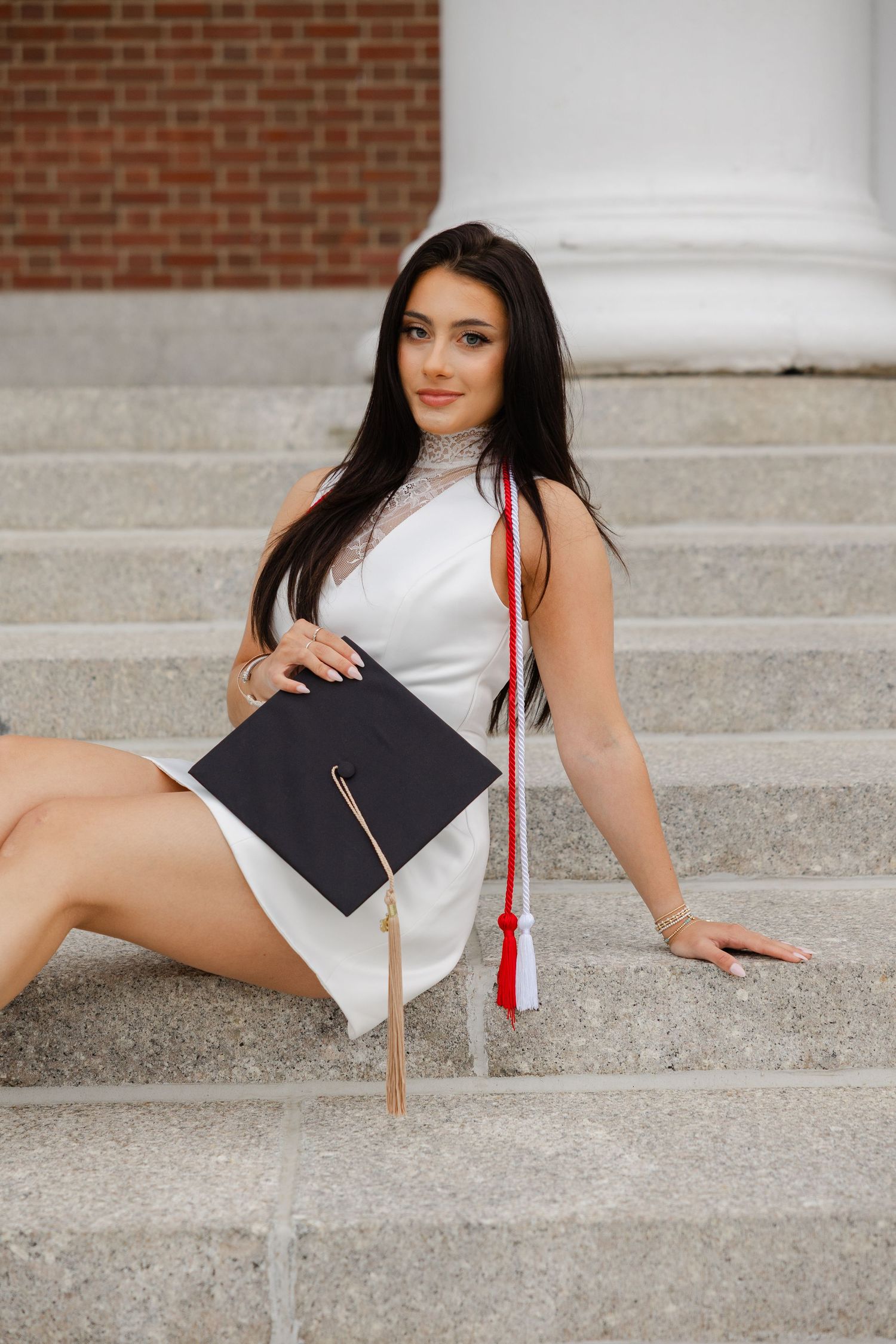 Young woman in white dress holding graduation cap and red tassel, sitting on stone steps outdoors.