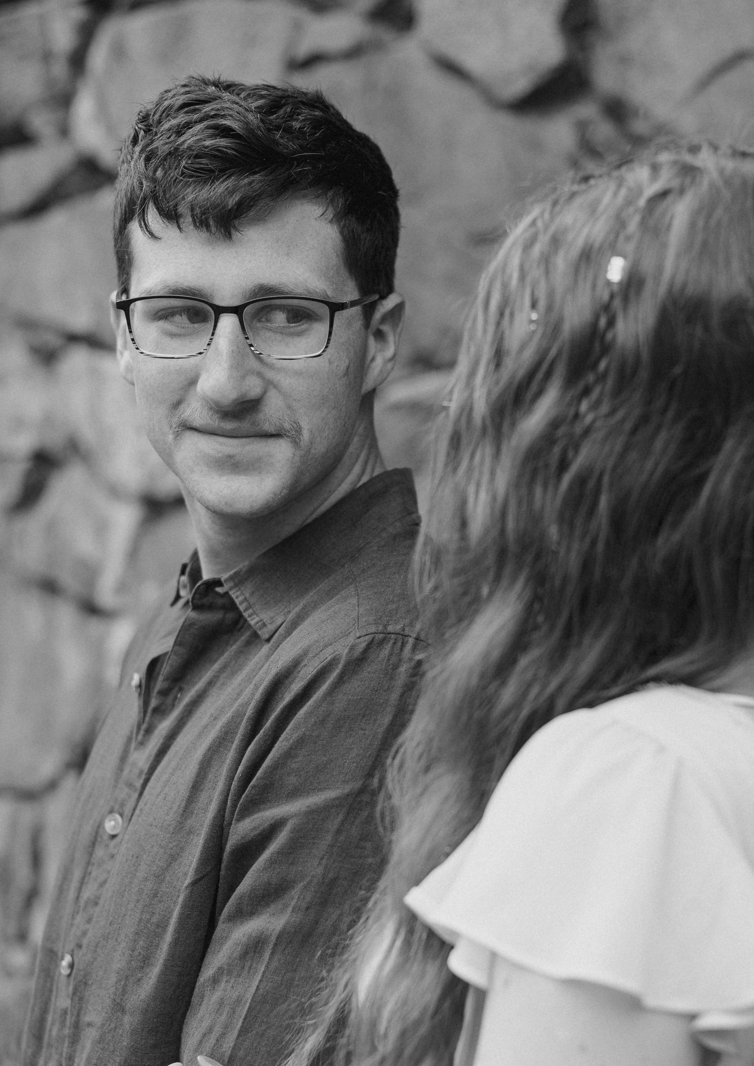 Black and white photo of a man with glasses smiling at a woman with long hair outdoors near a stone wall.