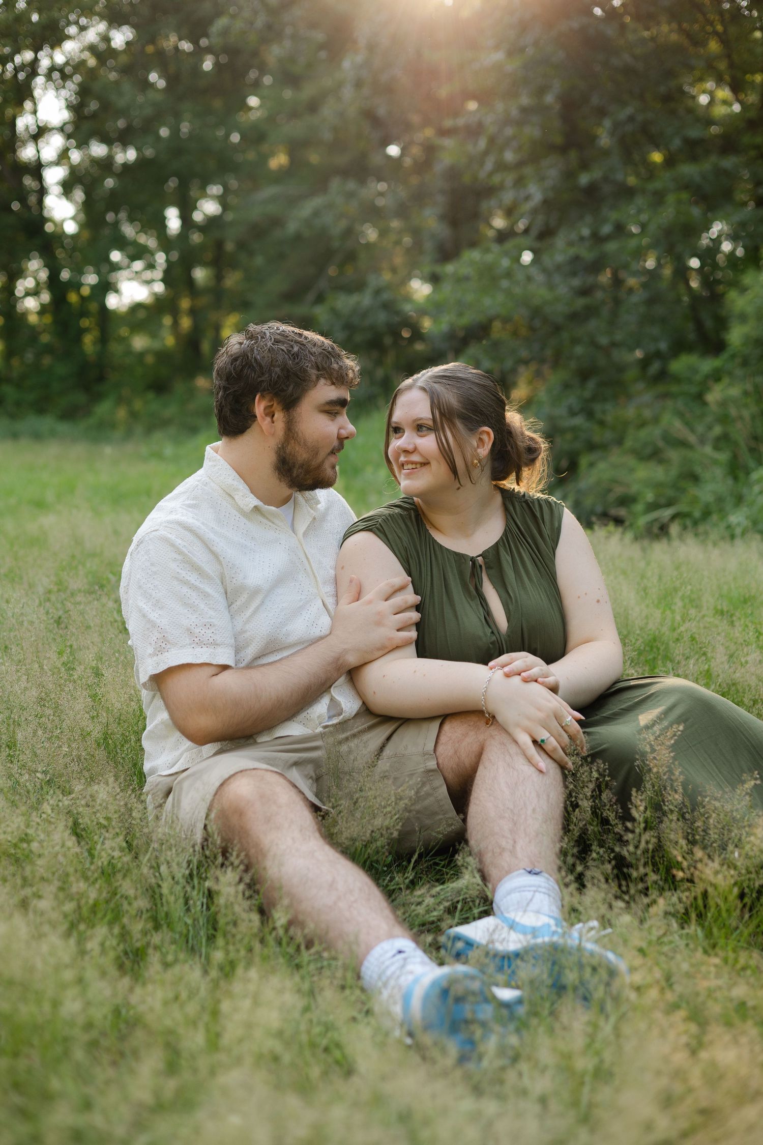 A couple sits together in a sunlit grassy field, smiling at each other, surrounded by lush green trees.