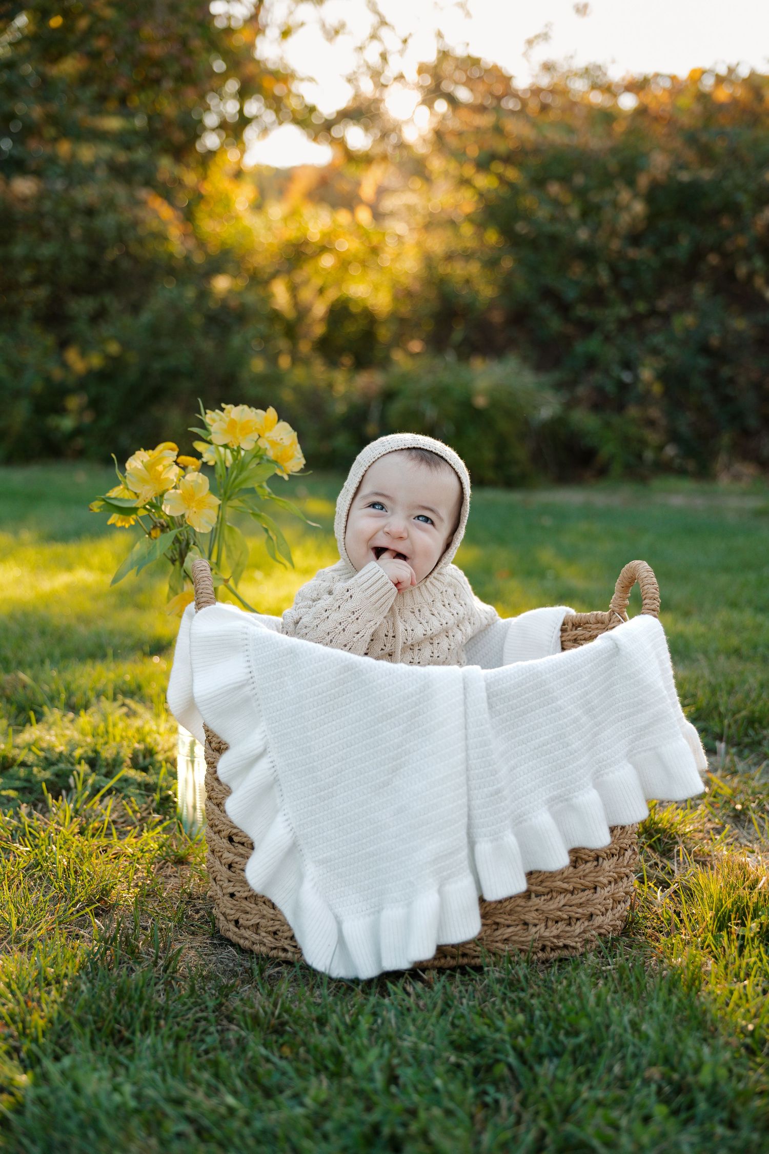 Adorable baby in a bonnet sitting in a wicker basket outdoors, holding yellow flowers, wrapped in a white ruffled blanket.