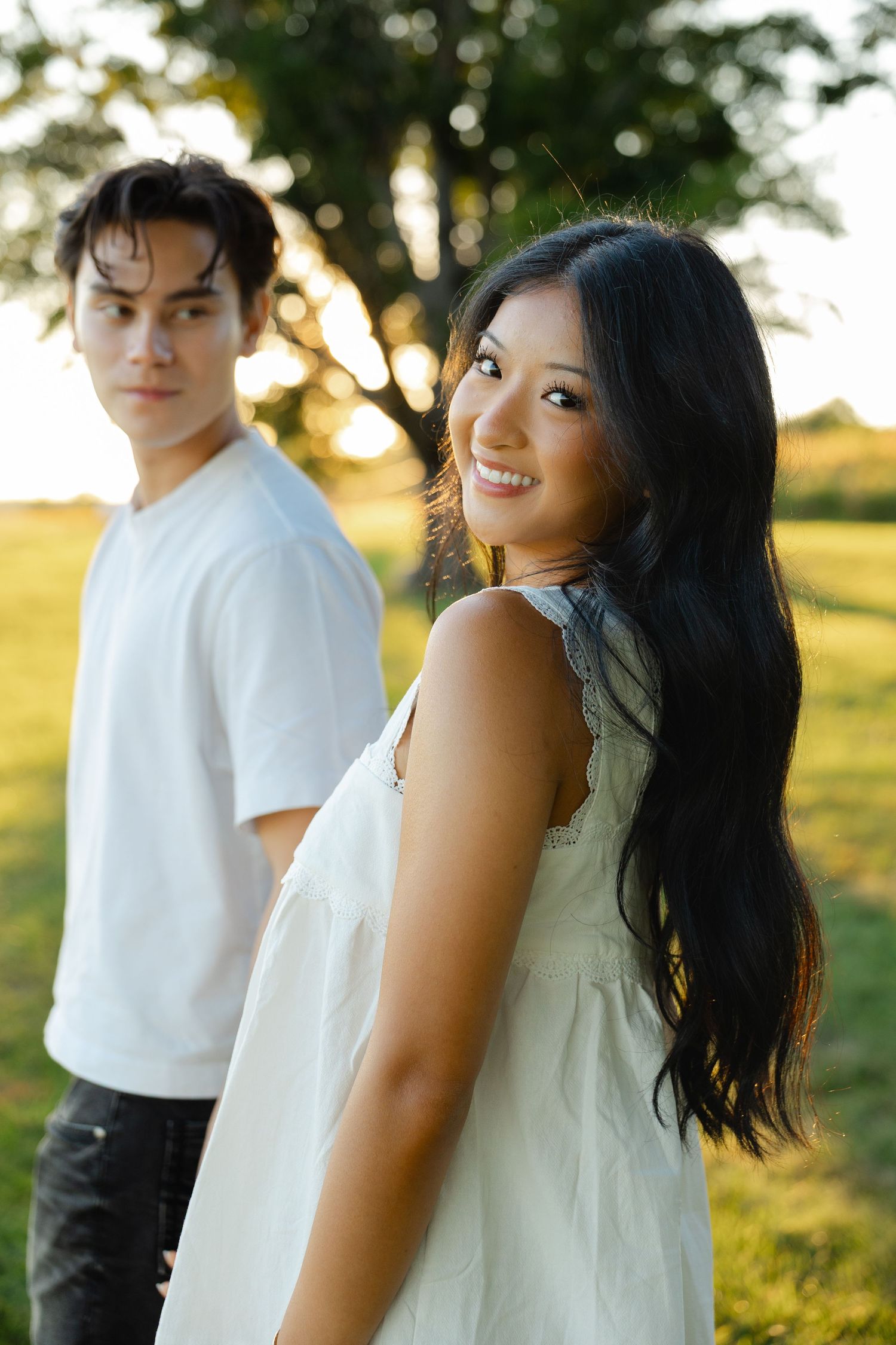 A smiling young Asian woman in a white dress turns back while a young man in a white tee stands behind her in a park.
