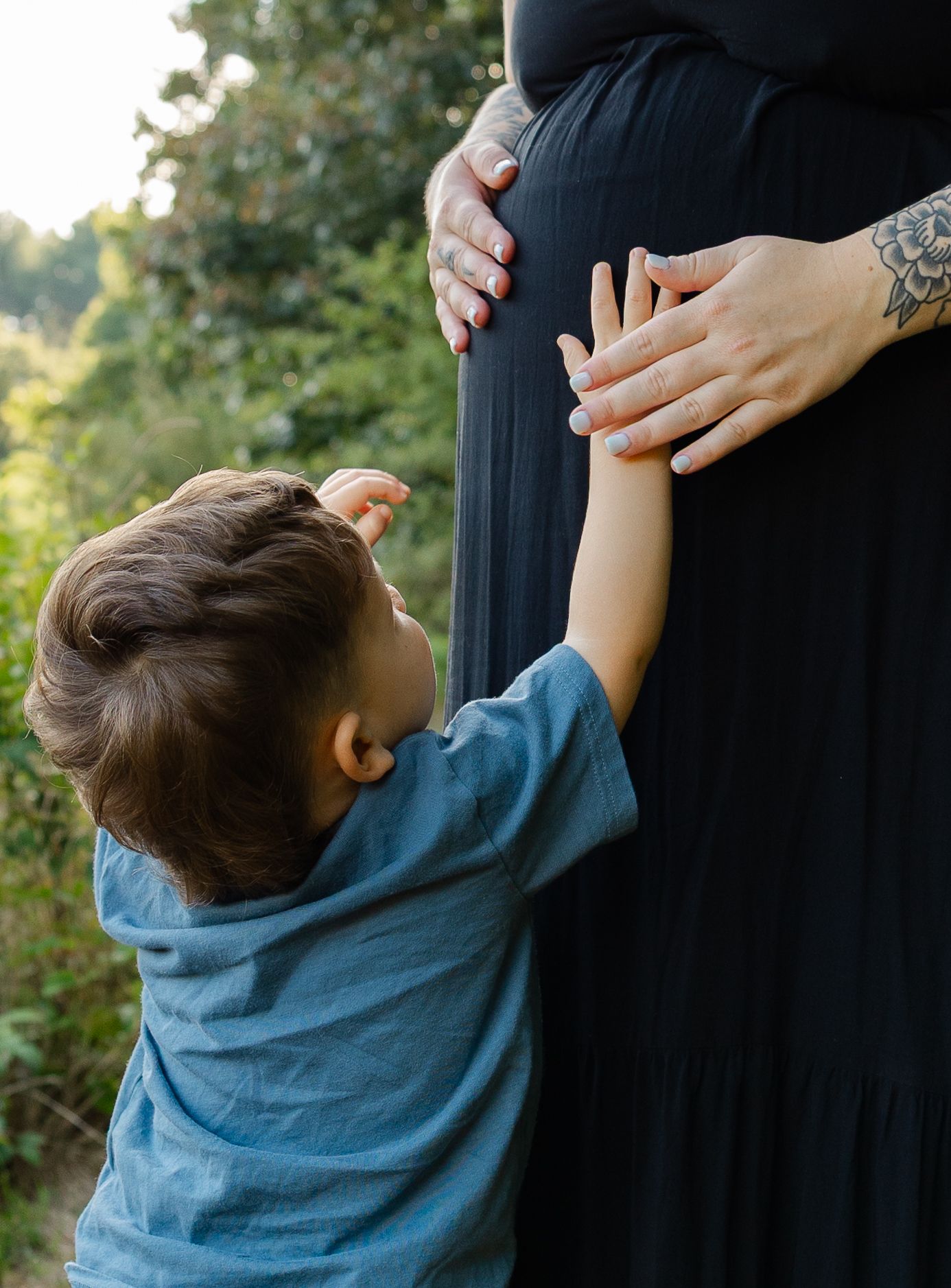A young toddler boy touches a pregnant woman's belly outdoors, wearing a blue shirt near green trees.