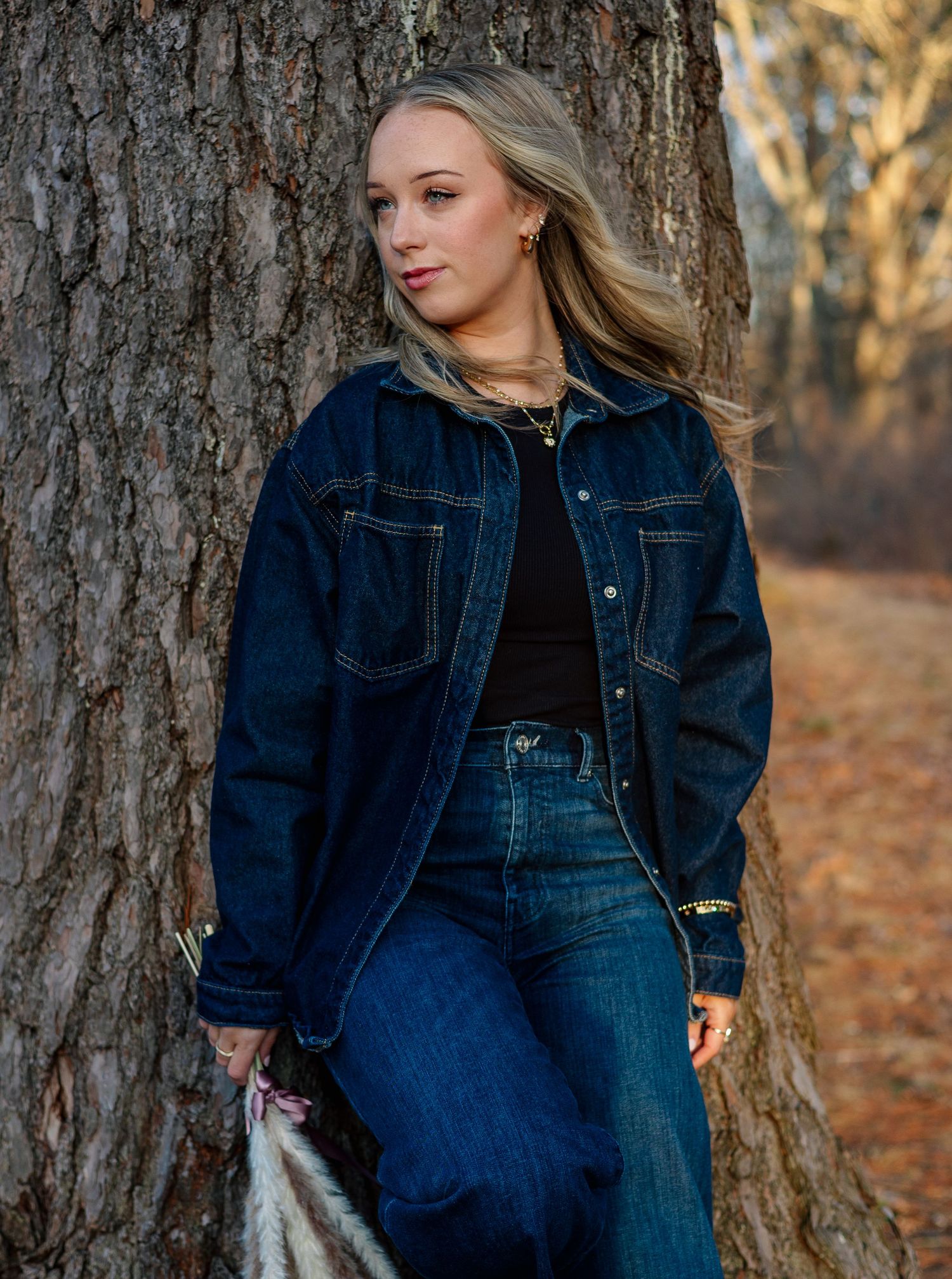 Young blonde woman in dark denim jacket leaning against tree trunk in wooded outdoor setting.