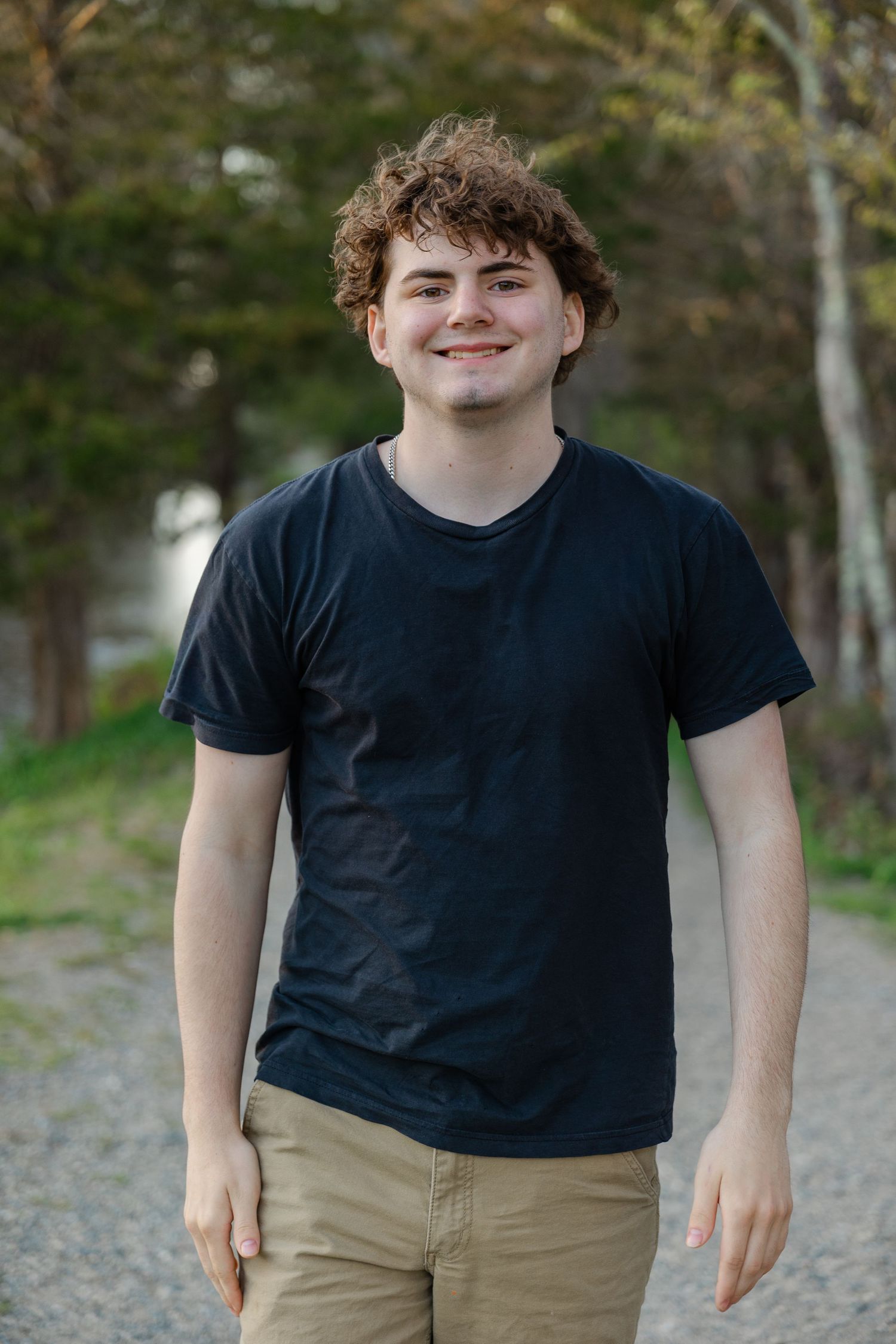 Young man with curly hair wearing a black t-shirt and khaki pants smiling outdoors on a gravel path.