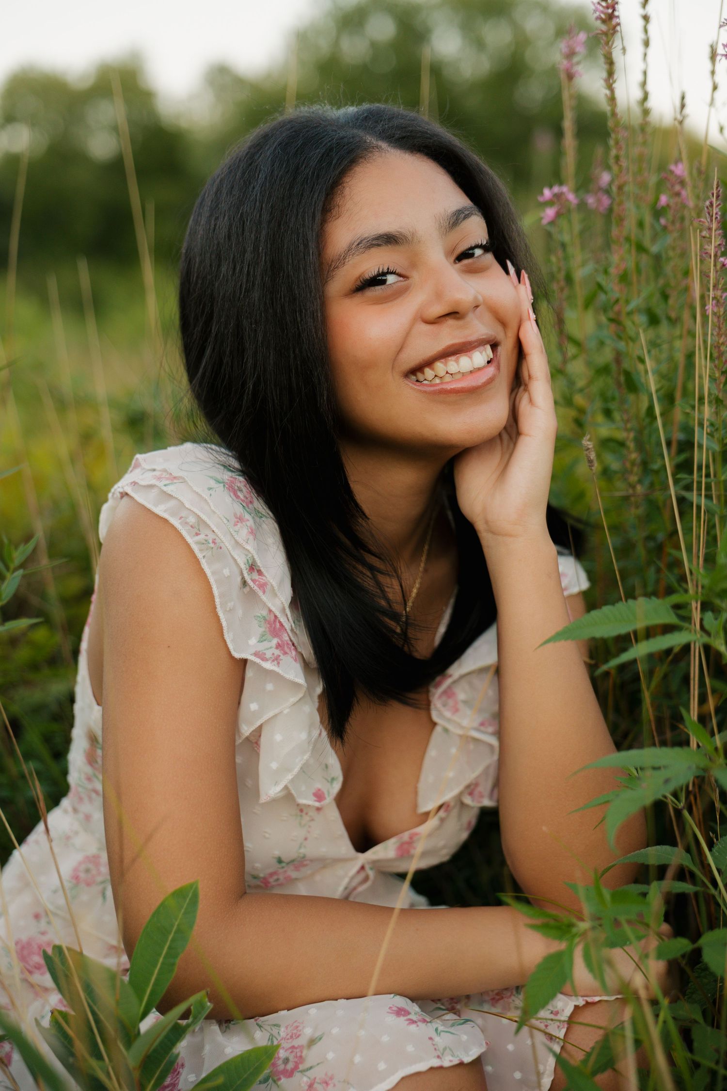 Young woman with dark hair smiles in a floral dress, sitting among wildflowers in a sunlit meadow.