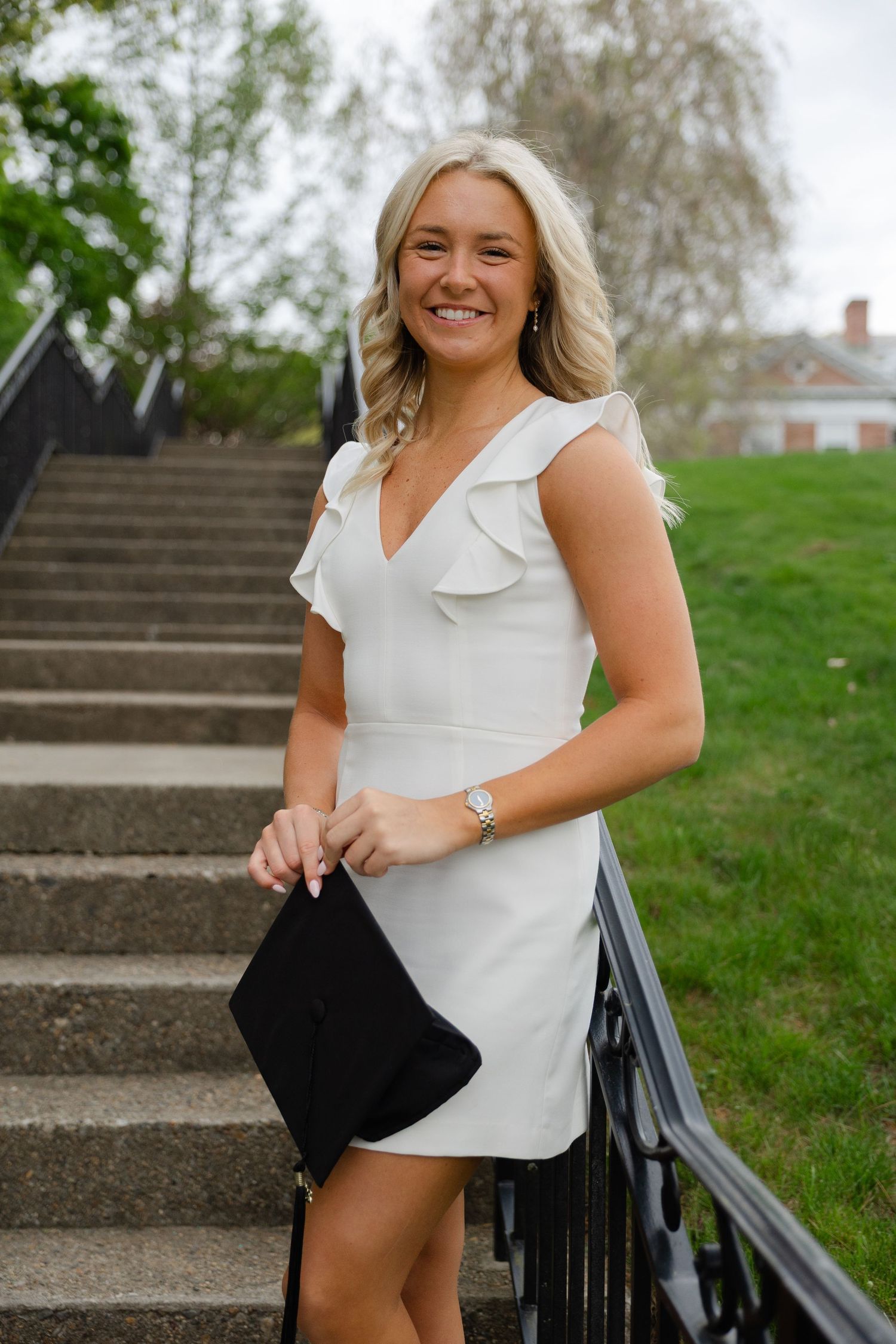 Smiling blonde woman in white ruffle dress holding black clutch on outdoor staircase.