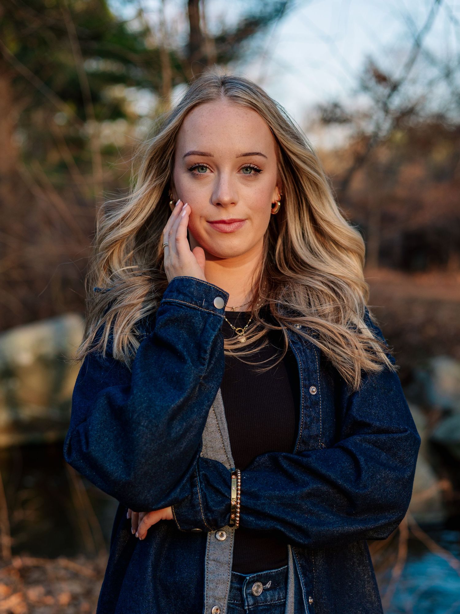 Young blonde woman in denim jacket posing outdoors near a creek in a natural wooded setting.