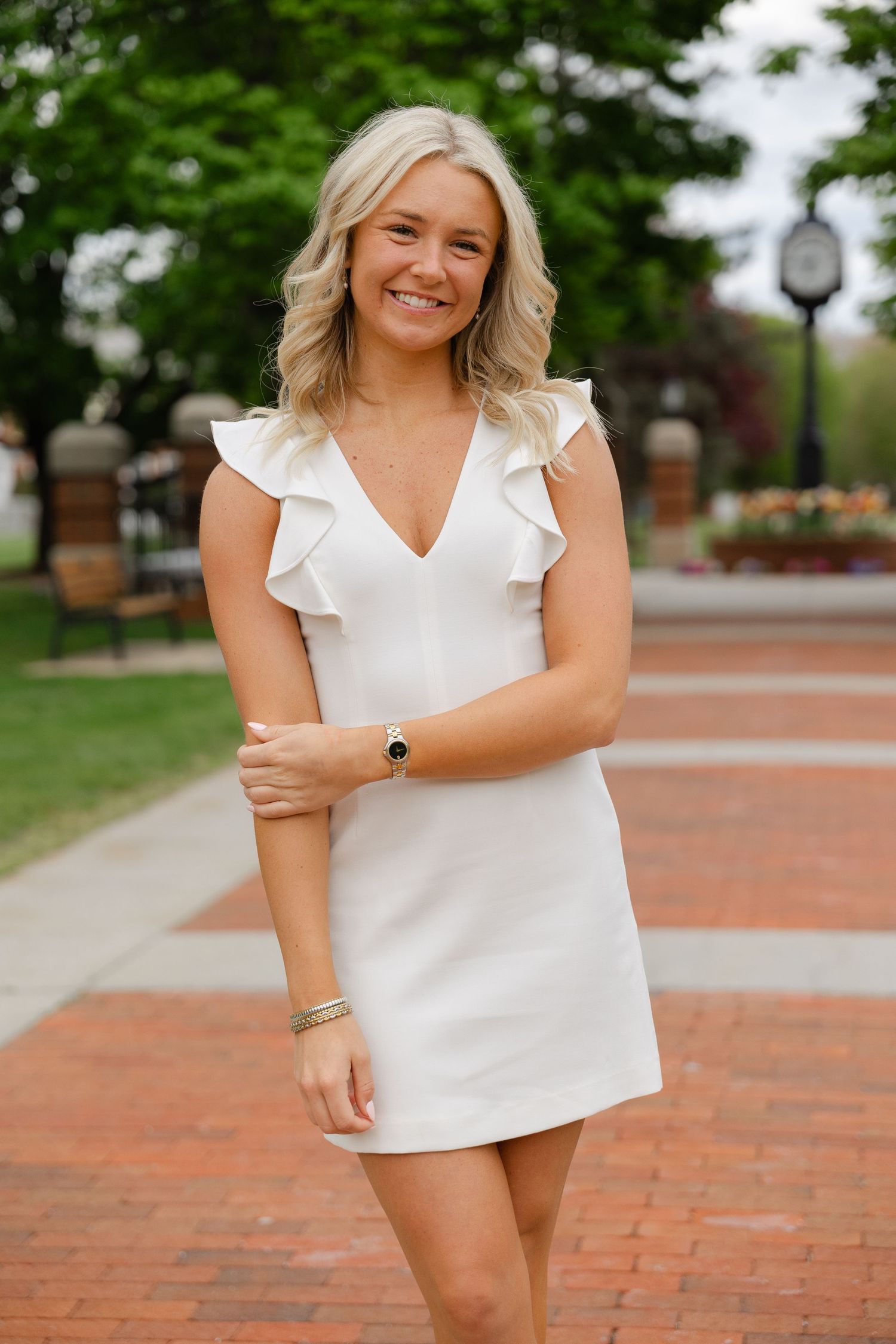 Young blonde woman smiling in white ruffle mini dress on brick walkway with green trees in background.