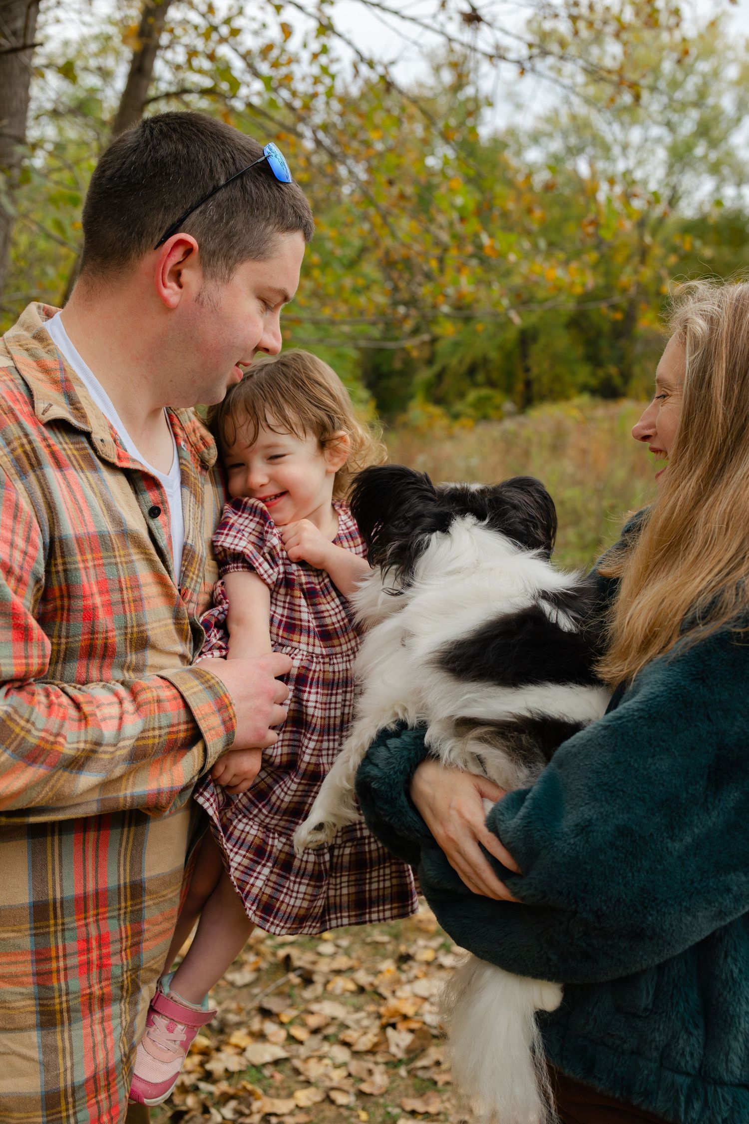 Family with toddler and black-white dog enjoying autumn outdoors, surrounded by colorful fall foliage.