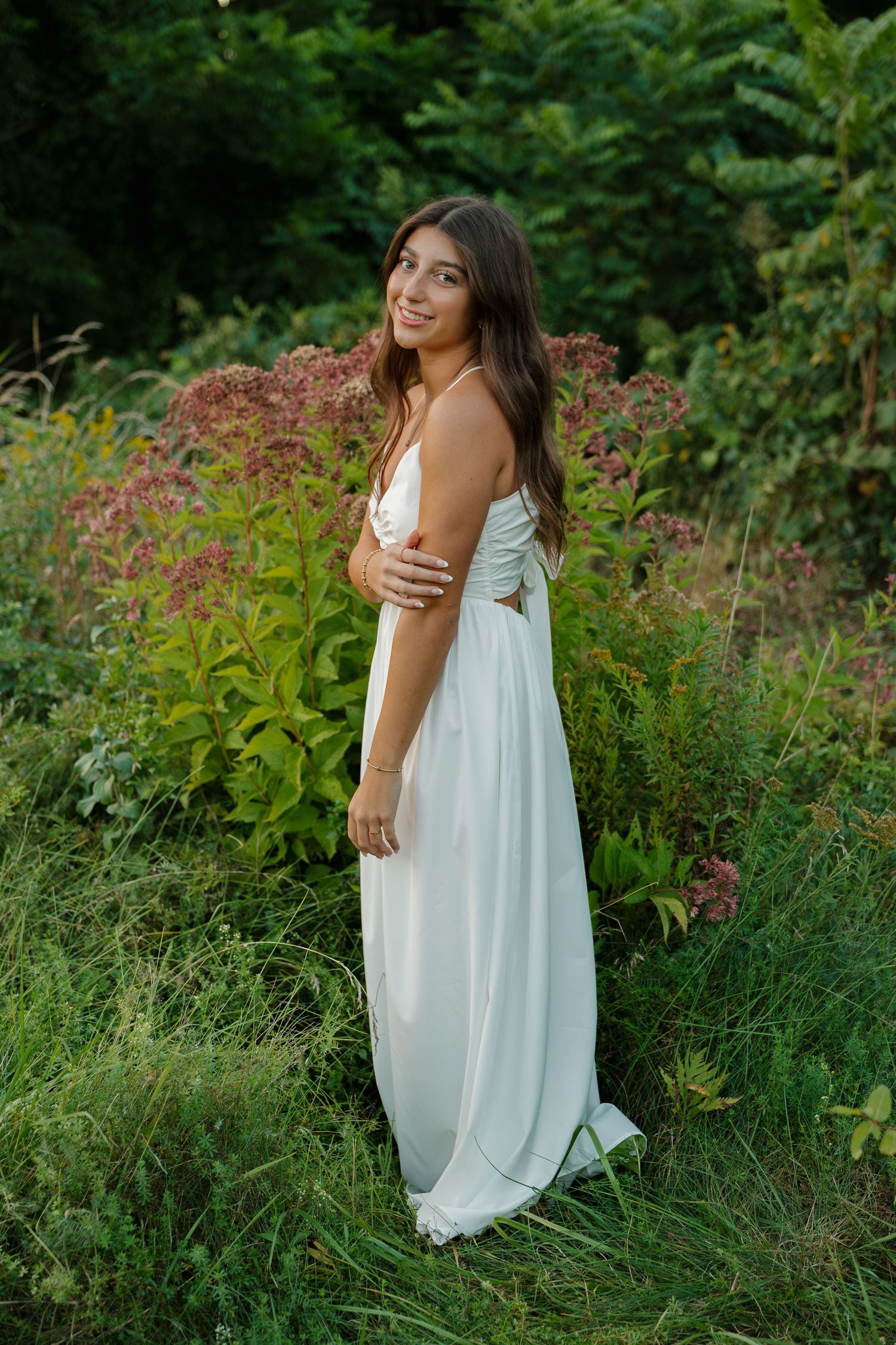 Young woman in a white maxi dress standing in a lush green garden with wildflowers, smiling over her shoulder.
