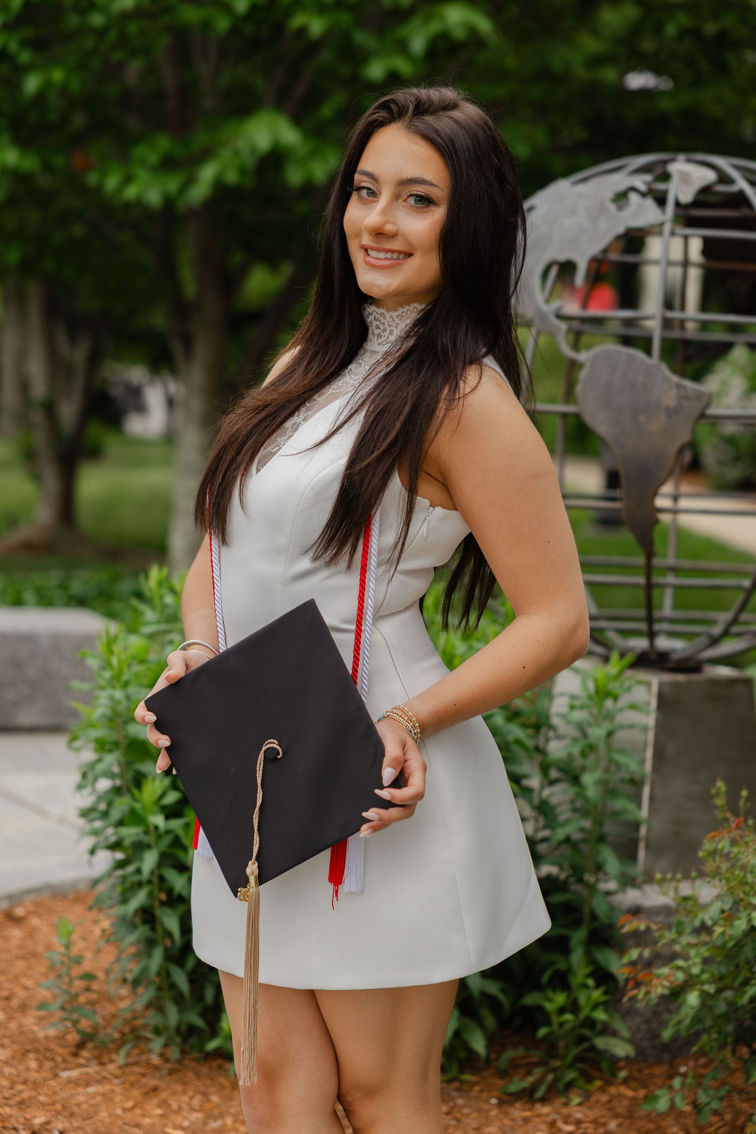 Young woman in white dress holding graduation cap smiles outdoors near globe sculpture on campus.