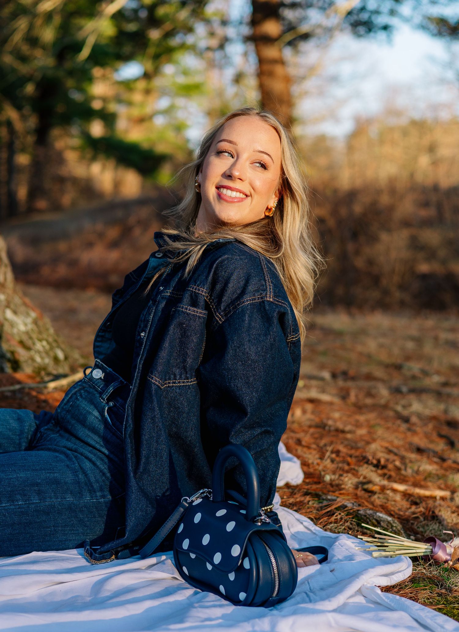 Smiling blonde woman in denim jacket sitting on white blanket outdoors with polka dot bag and flowers.