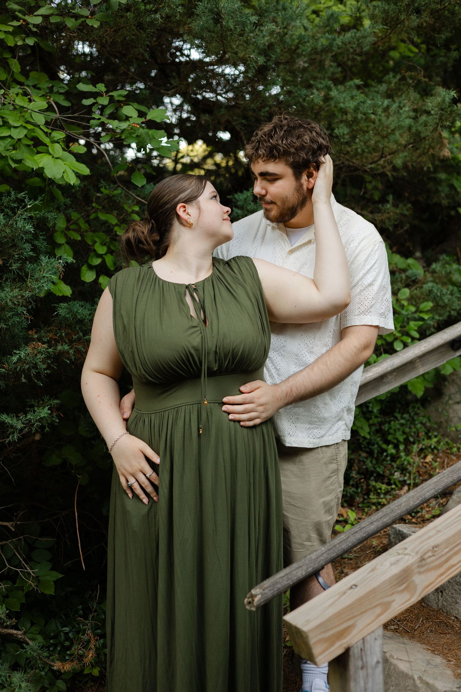 Pregnant couple in green dress and white shirt share tender moment on wooden steps surrounded by lush greenery.