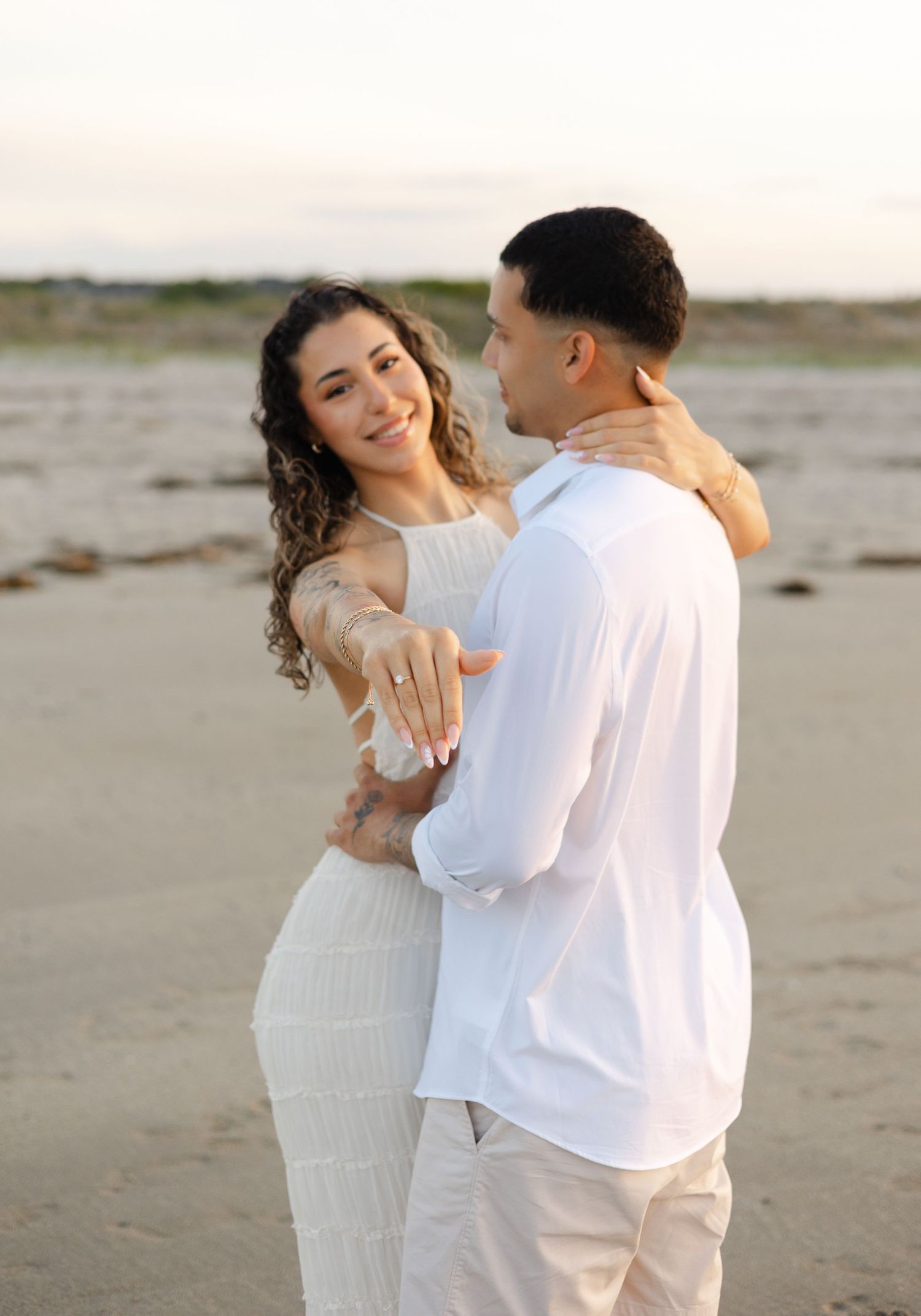 Couple in white outfits on beach, woman smiling and showing engagement ring, man facing away, sunset backdrop.