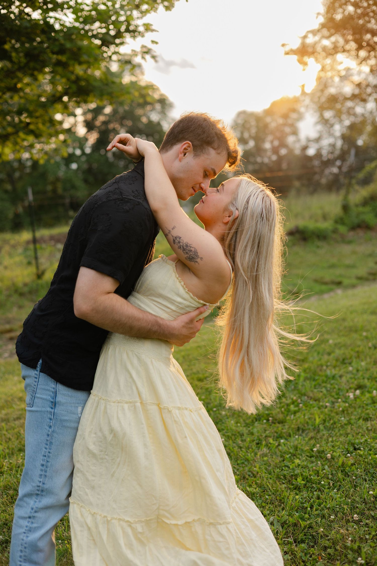 Couple nearly kissing at golden hour, woman in yellow dress and man in black shirt embracing outdoors.
