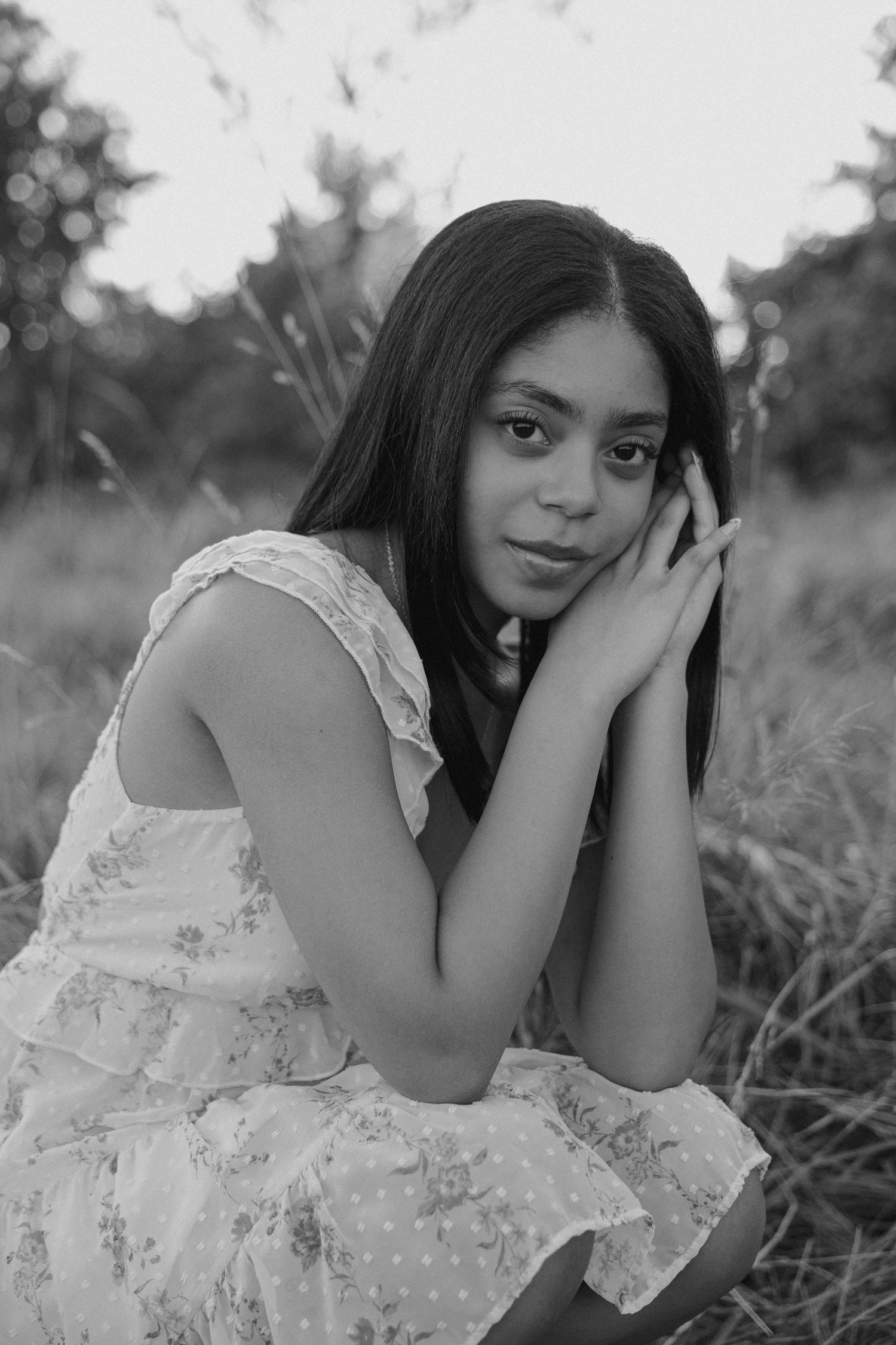Black and white portrait of a young woman in a floral dress sitting in a field, resting her face on her hands.