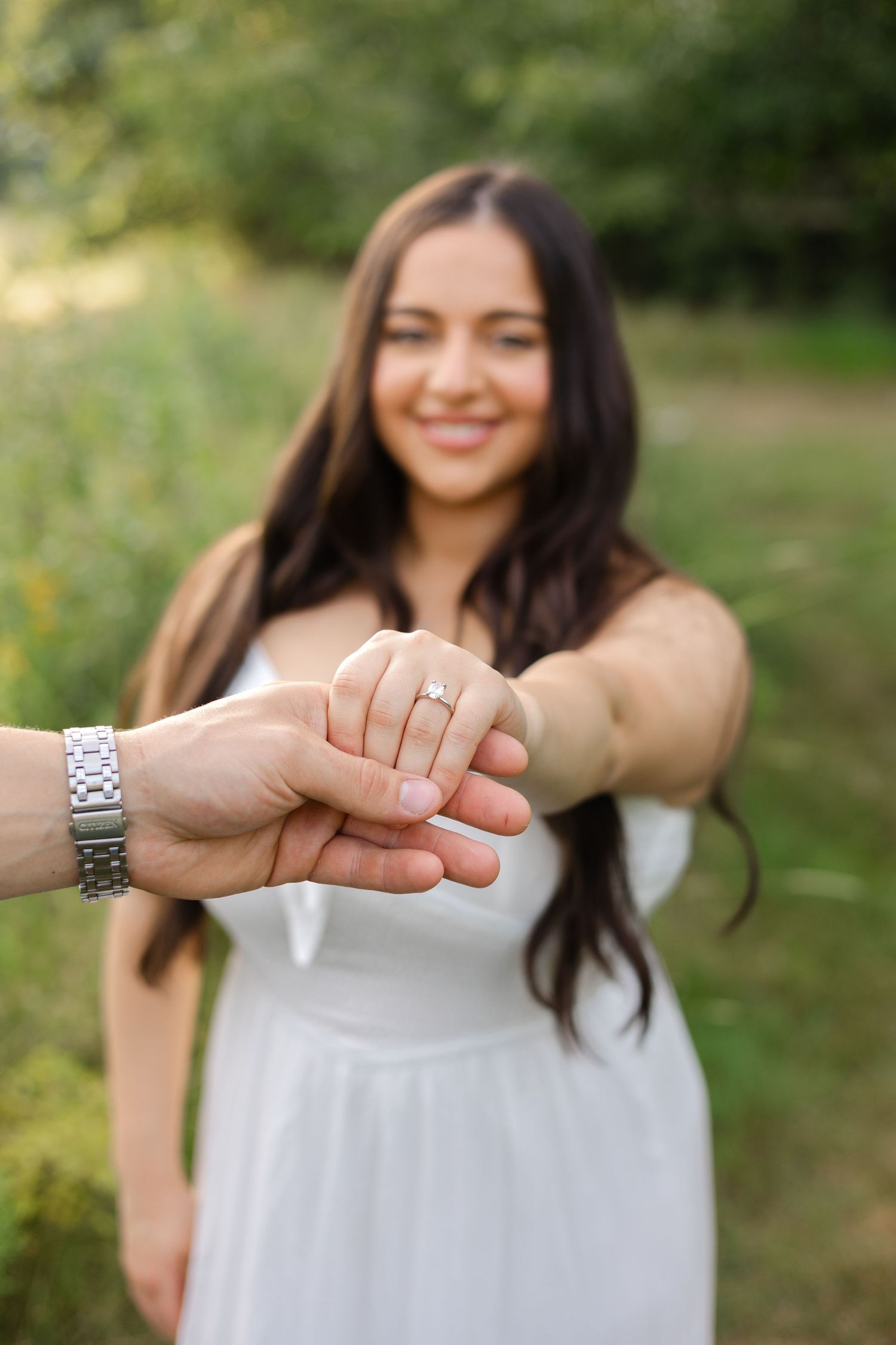 Smiling brunette woman in white dress showing off engagement ring while holding hands with partner outdoors.